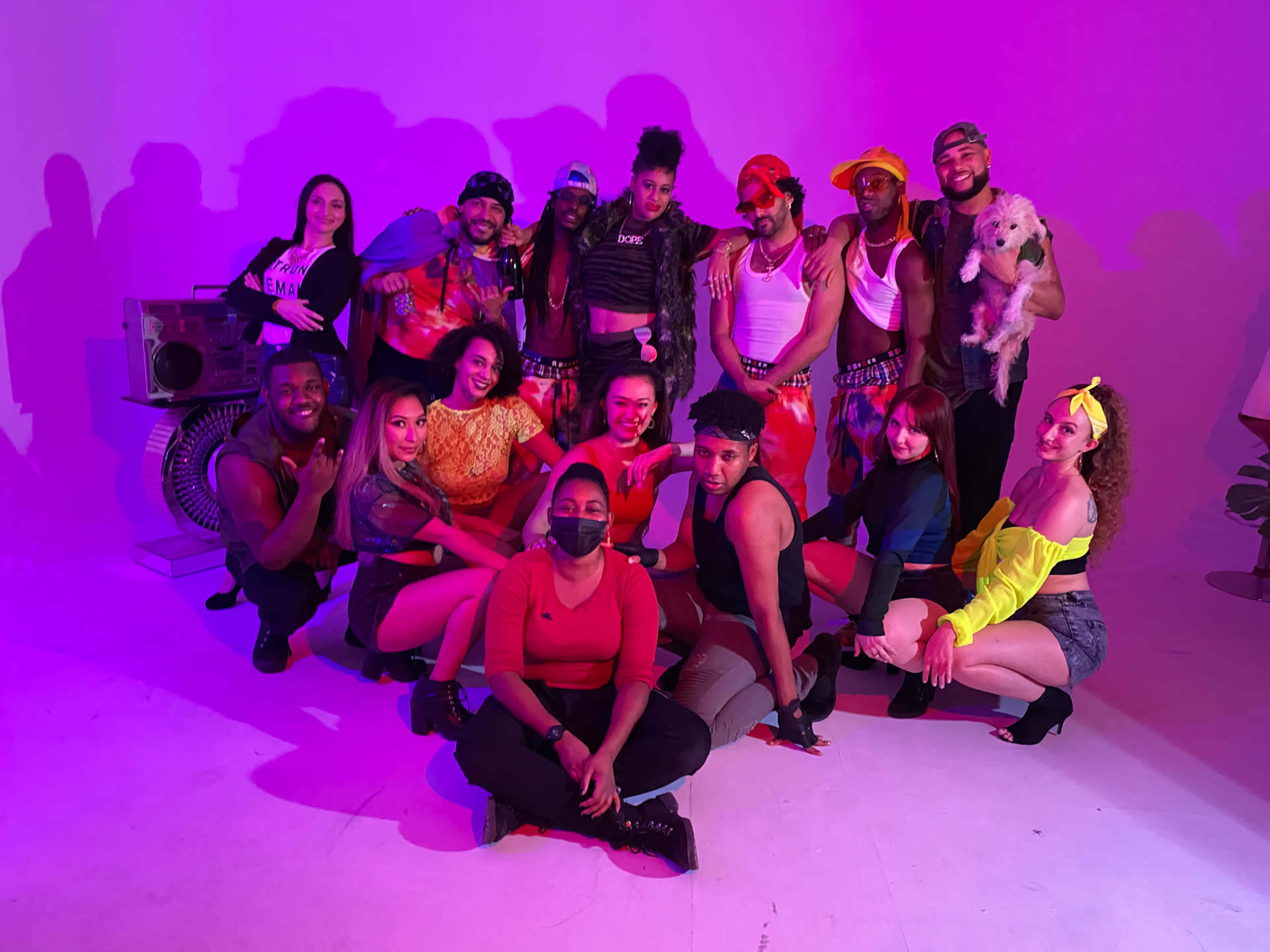 A diverse group of people poses together in front of a purple-lit background, some holding a small dog and surrounded by retro props.