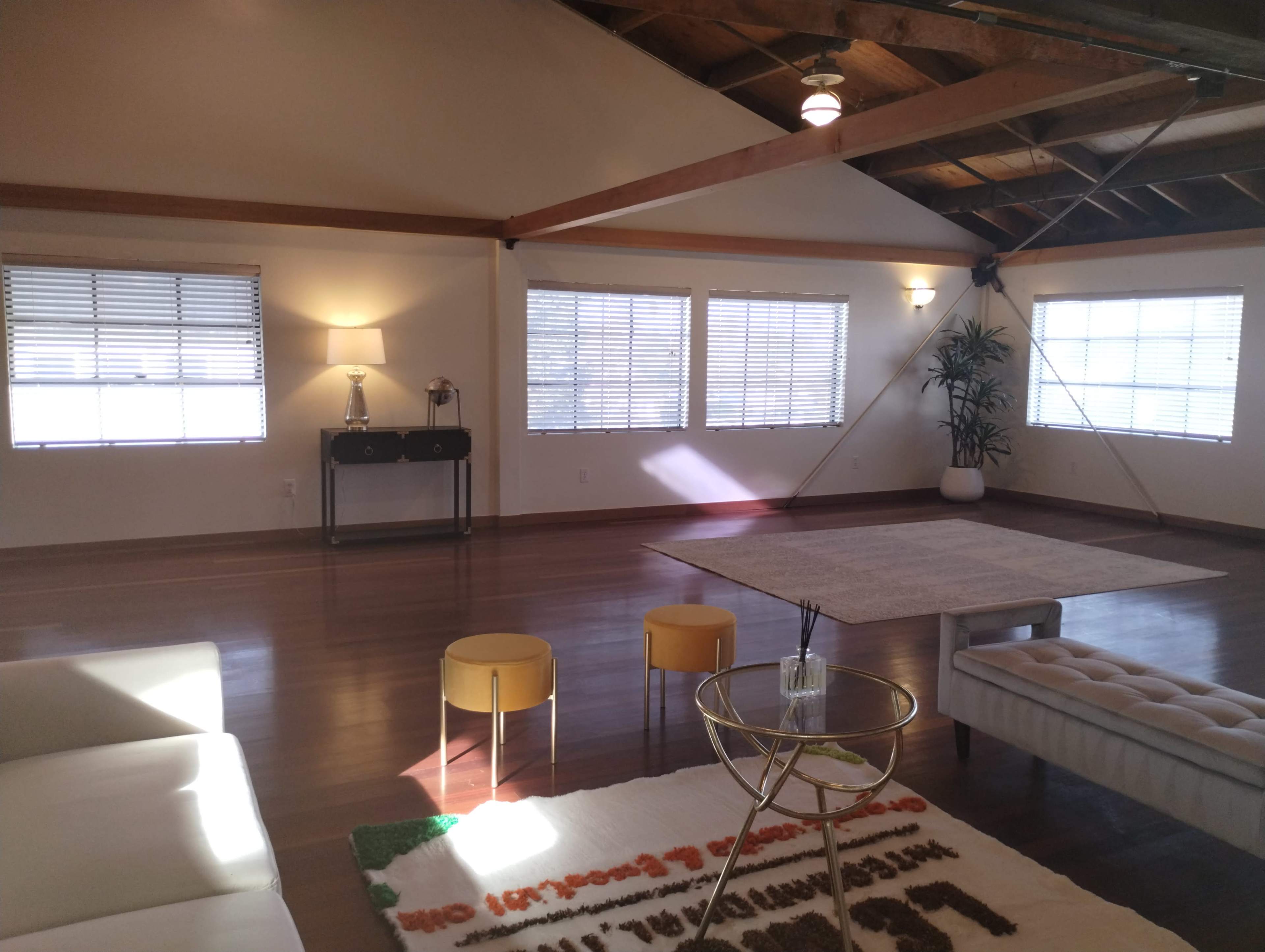 A spacious, well-lit room features large windows, light-colored walls, and wooden beams, furnished with a sofa, a coffee table, and small stools.