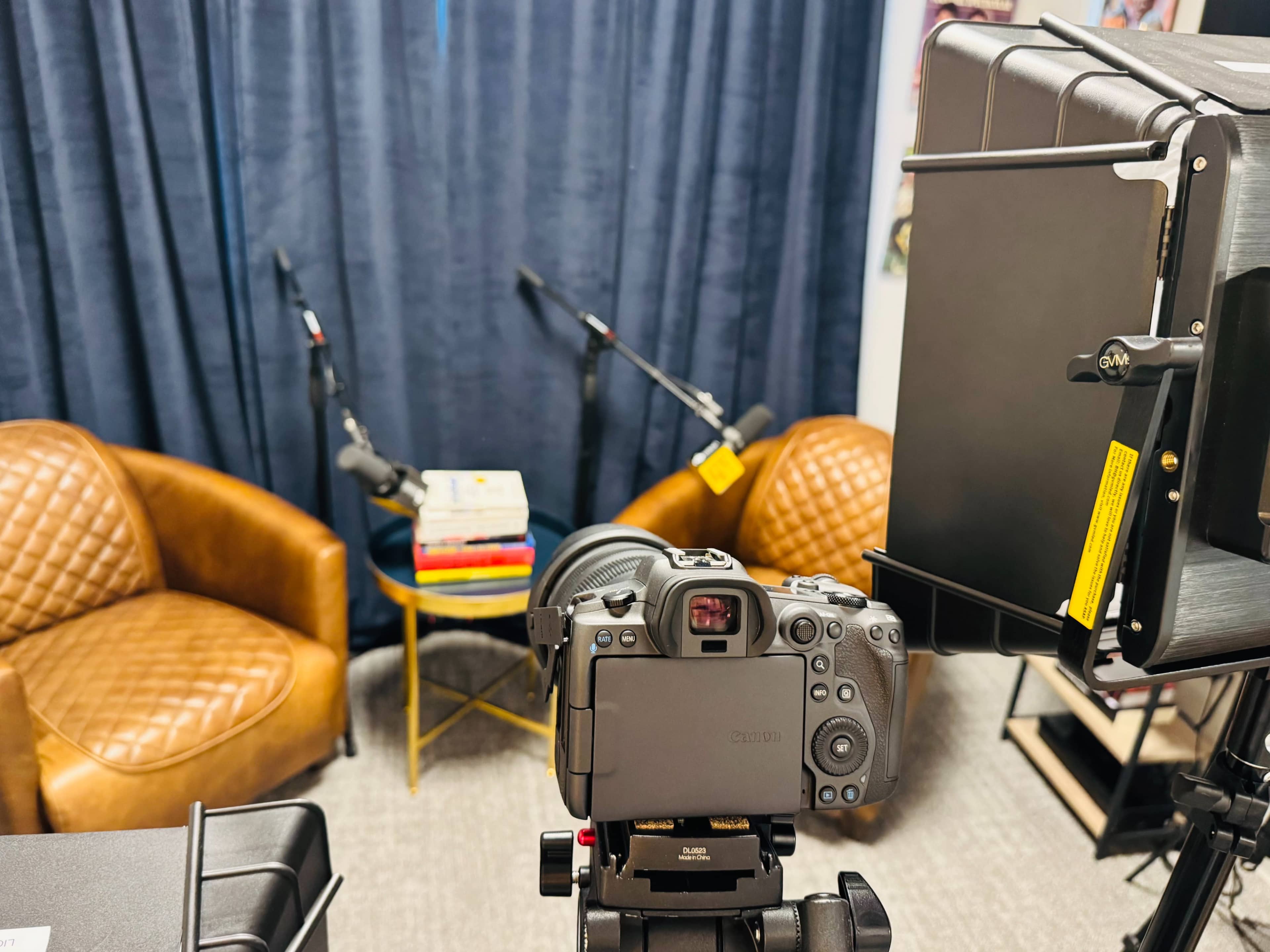 A camera is positioned in front of two brown chairs and a small table stacked with books, with microphones set up nearby against a backdrop of blue curtains.