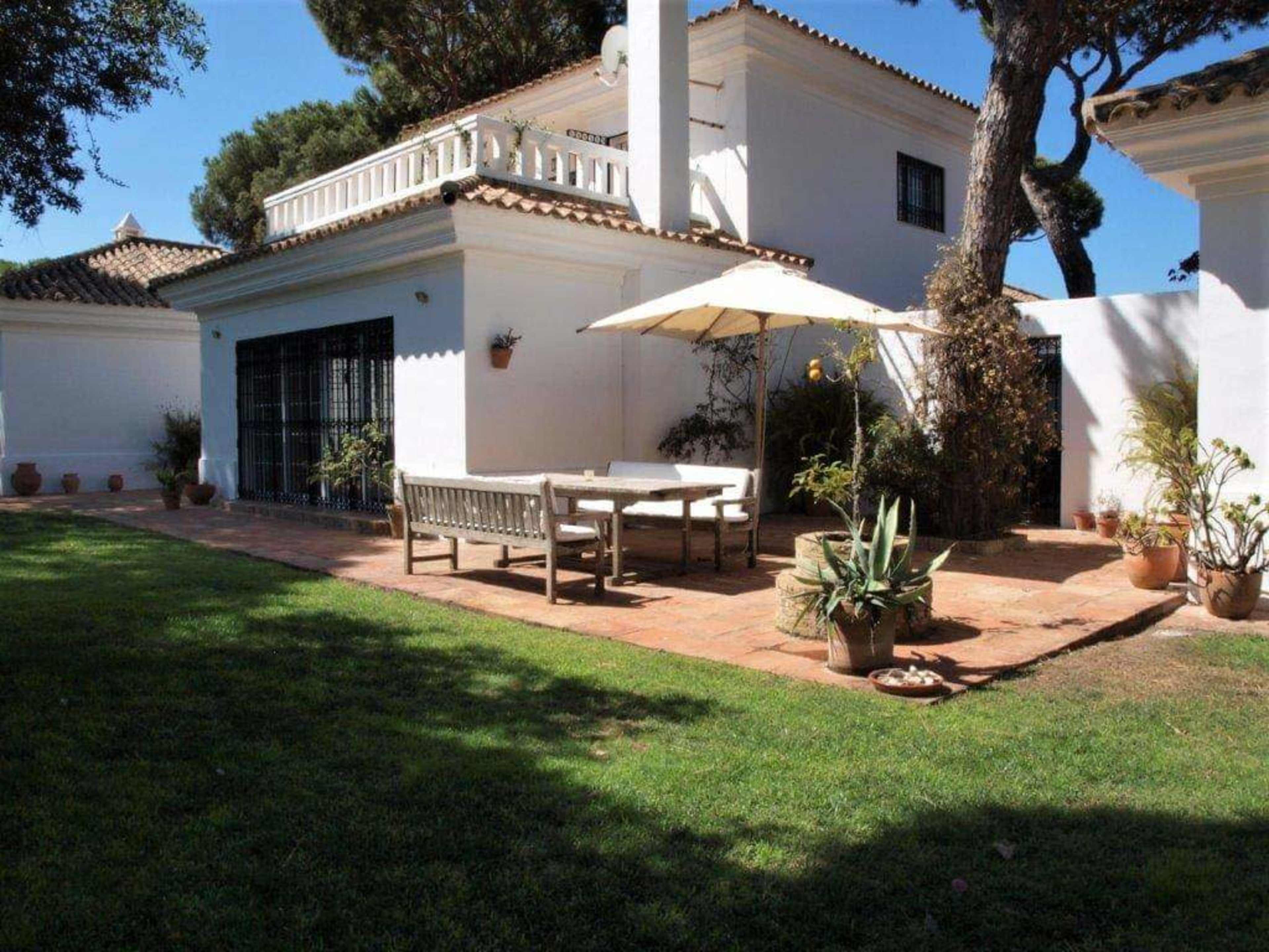 A patio area with a white table, chairs, and an umbrella is surrounded by potted plants near a white house.