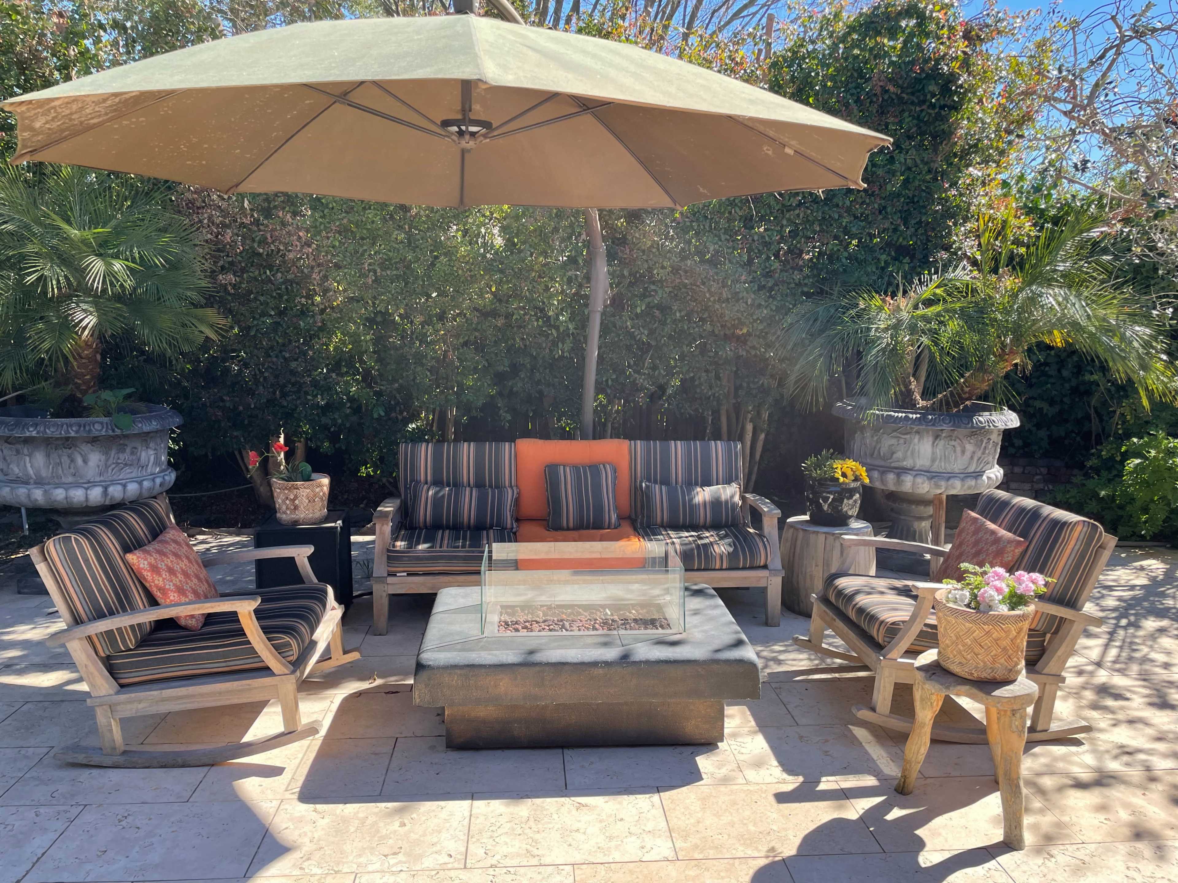A shaded outdoor seating area features a sofa and chairs arranged around a coffee table with a firepit, surrounded by decorative planters and palm trees.