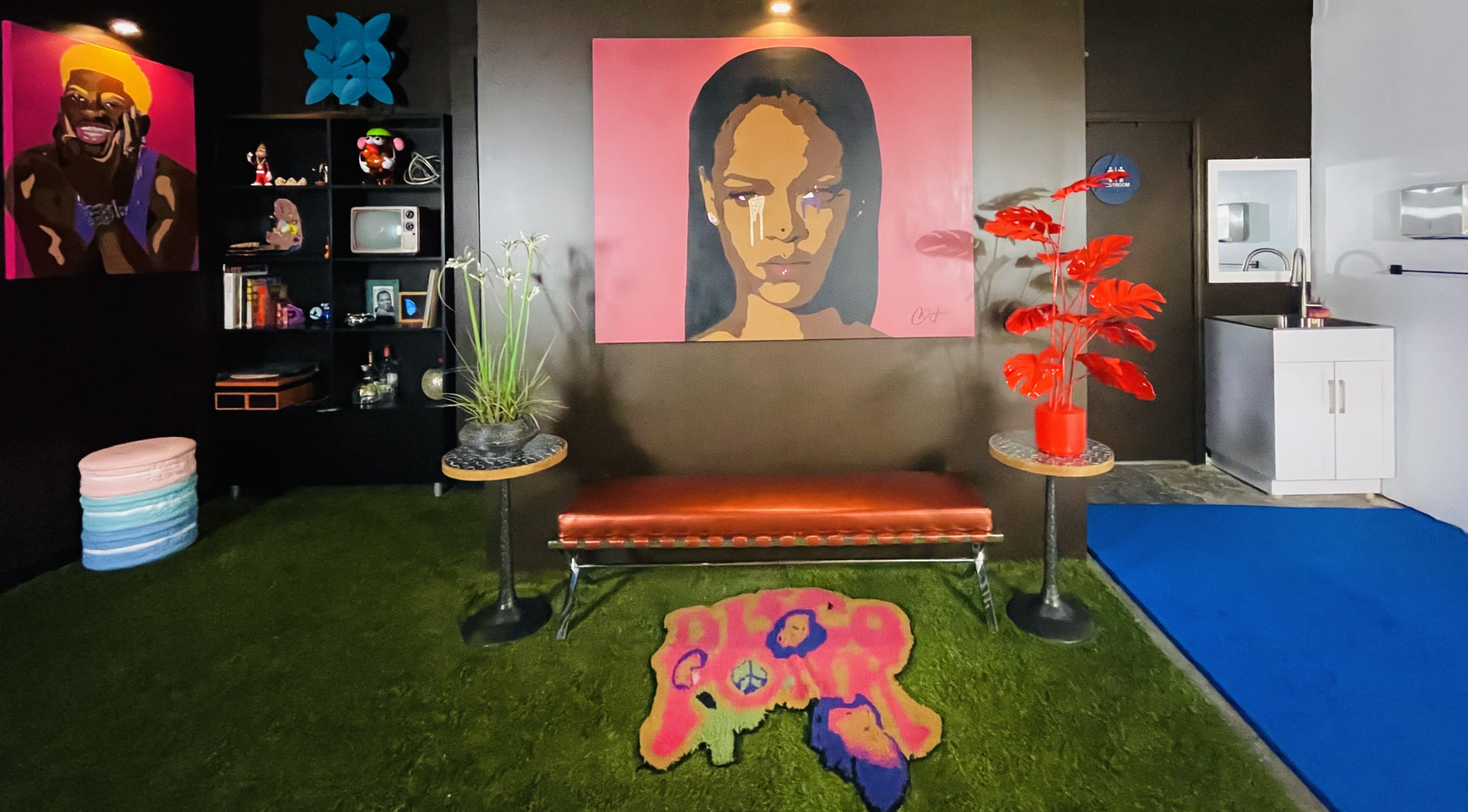 The image shows a colorful interior space featuring a large portrait on the wall, a bench, vibrant plants, and carpeted areas.