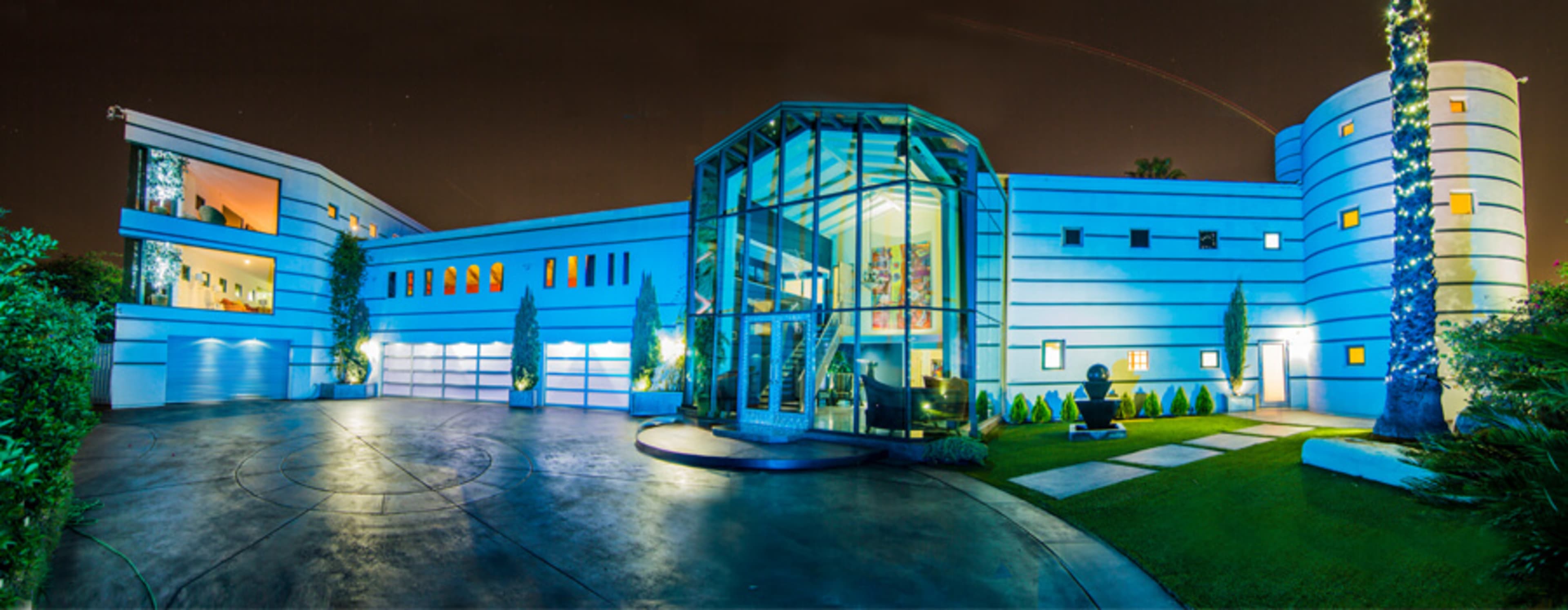 A modern, two-story house features a glass-fronted entryway and is illuminated in blue at night, surrounded by landscaped greenery and a curved driveway.