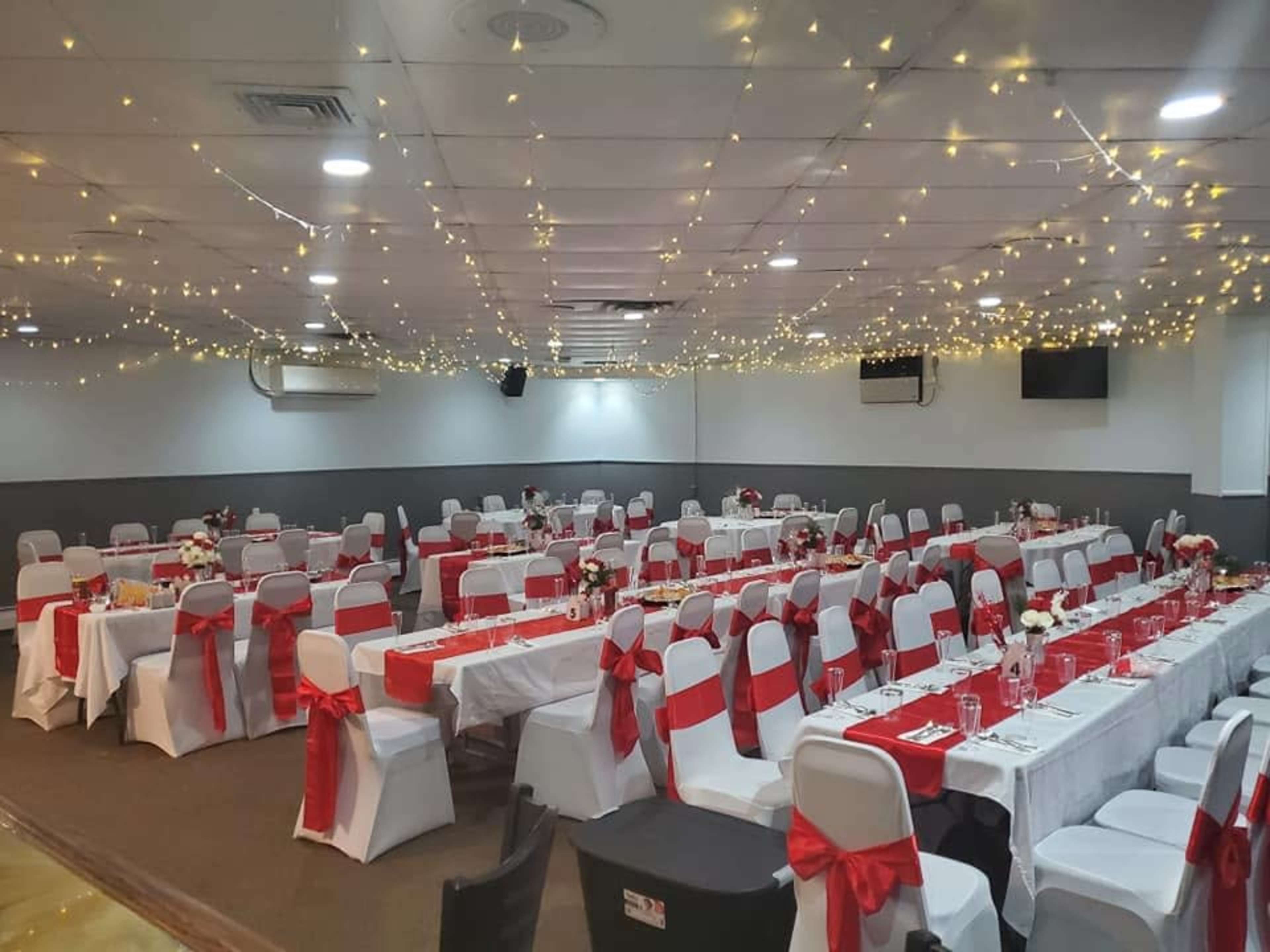 The image shows a banquet hall arranged for a formal event, with rows of tables covered in white cloths and decorated with red ribbons and floral centerpieces, and string lights hanging from the ceiling.