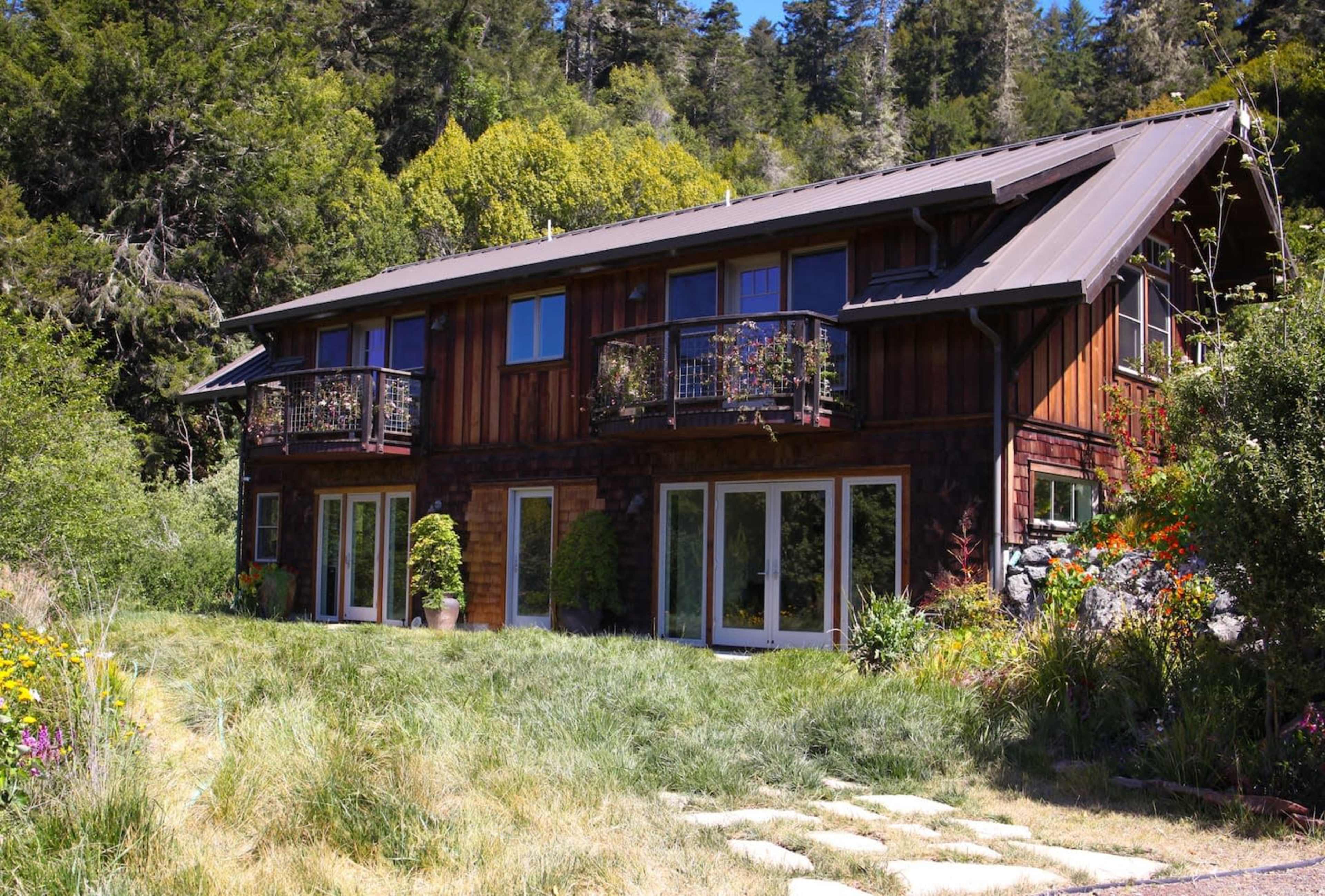 A two-story wooden house with balconies is surrounded by greenery and flowers.