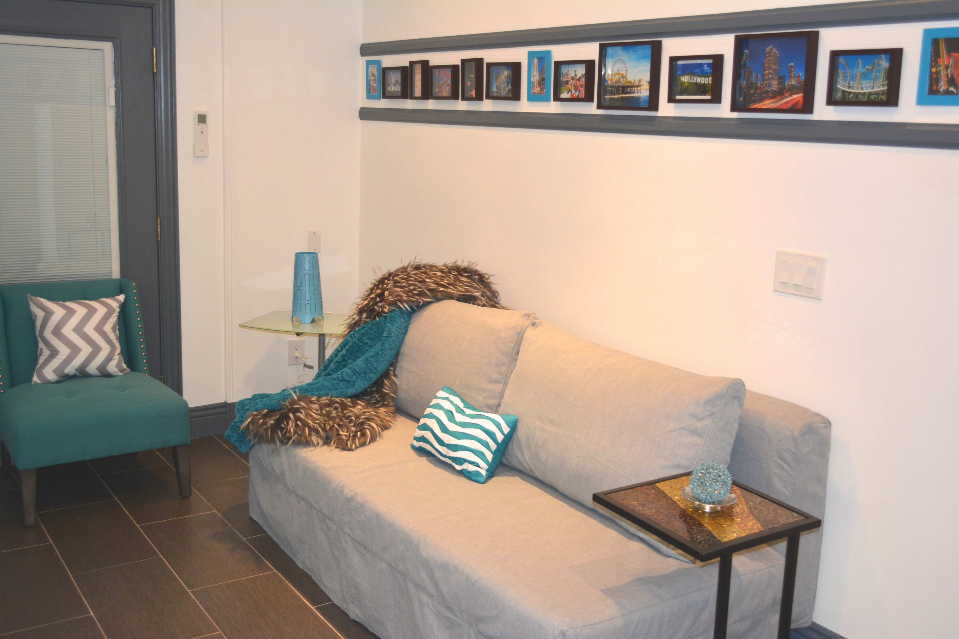 The image shows a living room with a gray sofa, a teal accent chair, a small glass side table, and a series of framed pictures mounted on the wall.