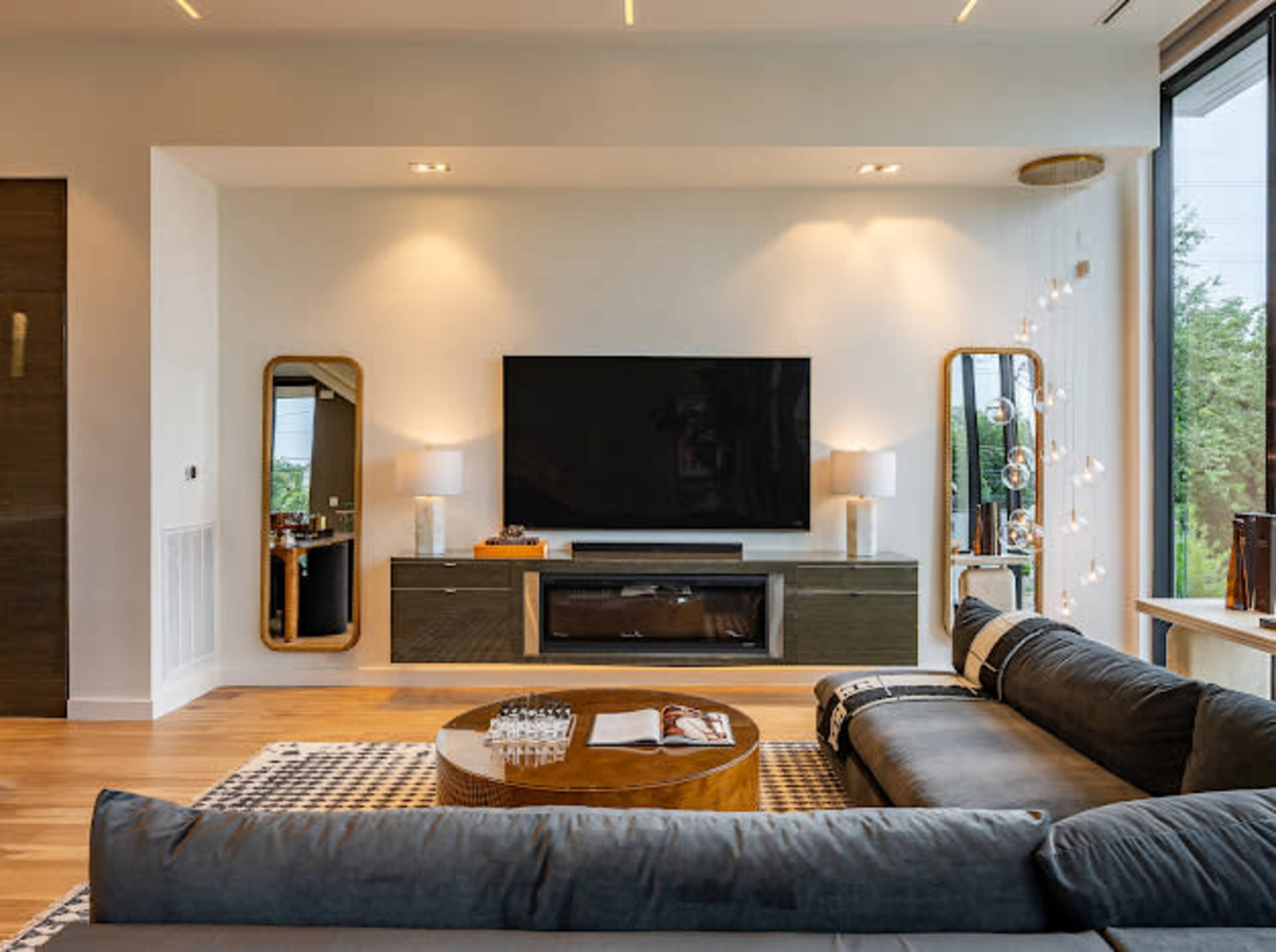 The image shows a modern living room featuring a large flat-screen TV mounted on a wall, a sectional sofa, a circular coffee table, and decorative mirrors on either side of the TV.