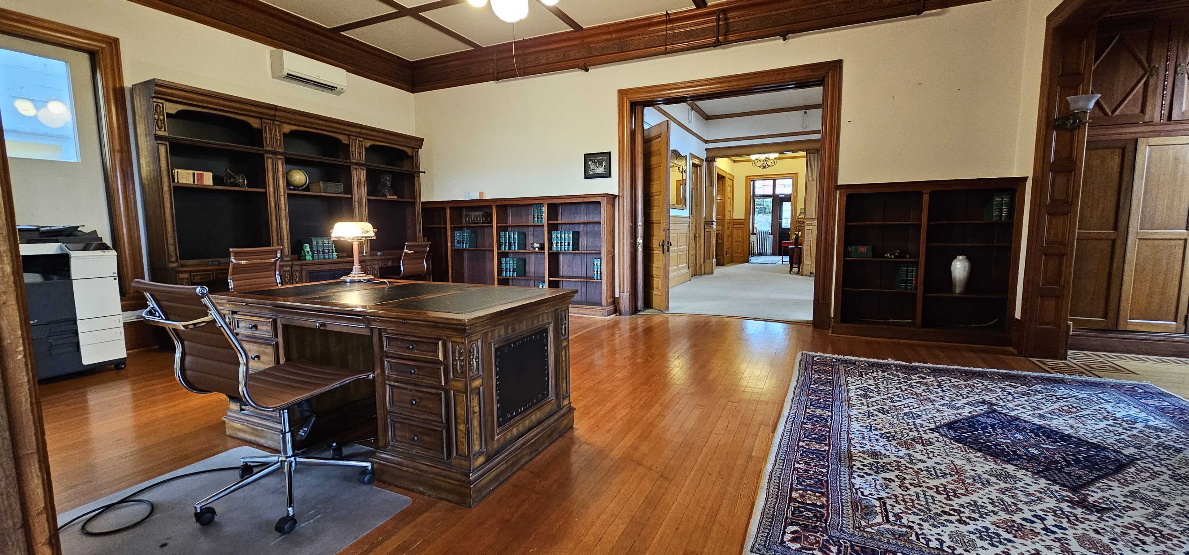 A wooden desk with a lamp in a spacious office featuring bookshelves and a doorway leading to another room.