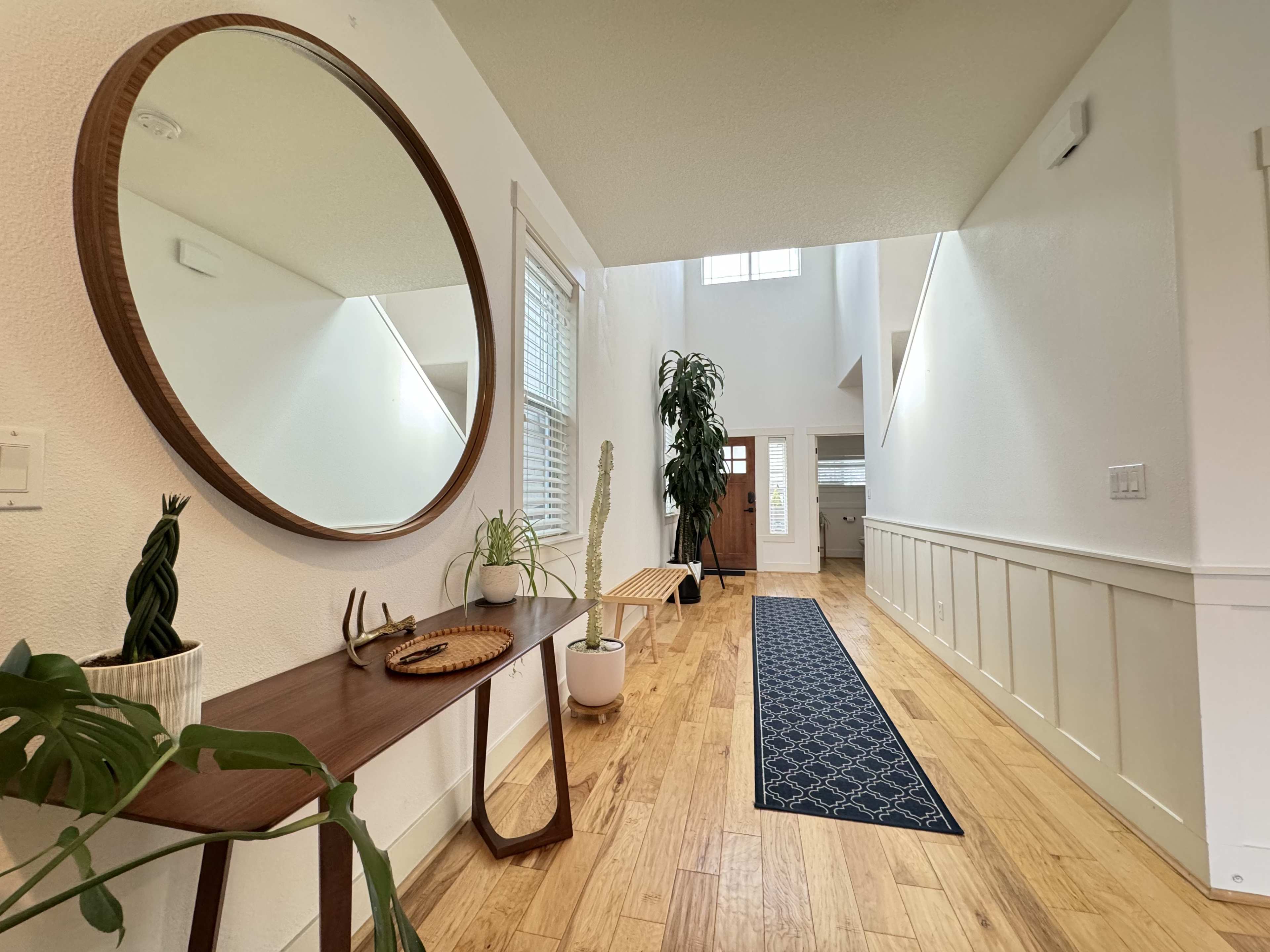 A bright hallway features a round mirror on a console table, a potted plant, and a long rug leading to a doorway at the far end.