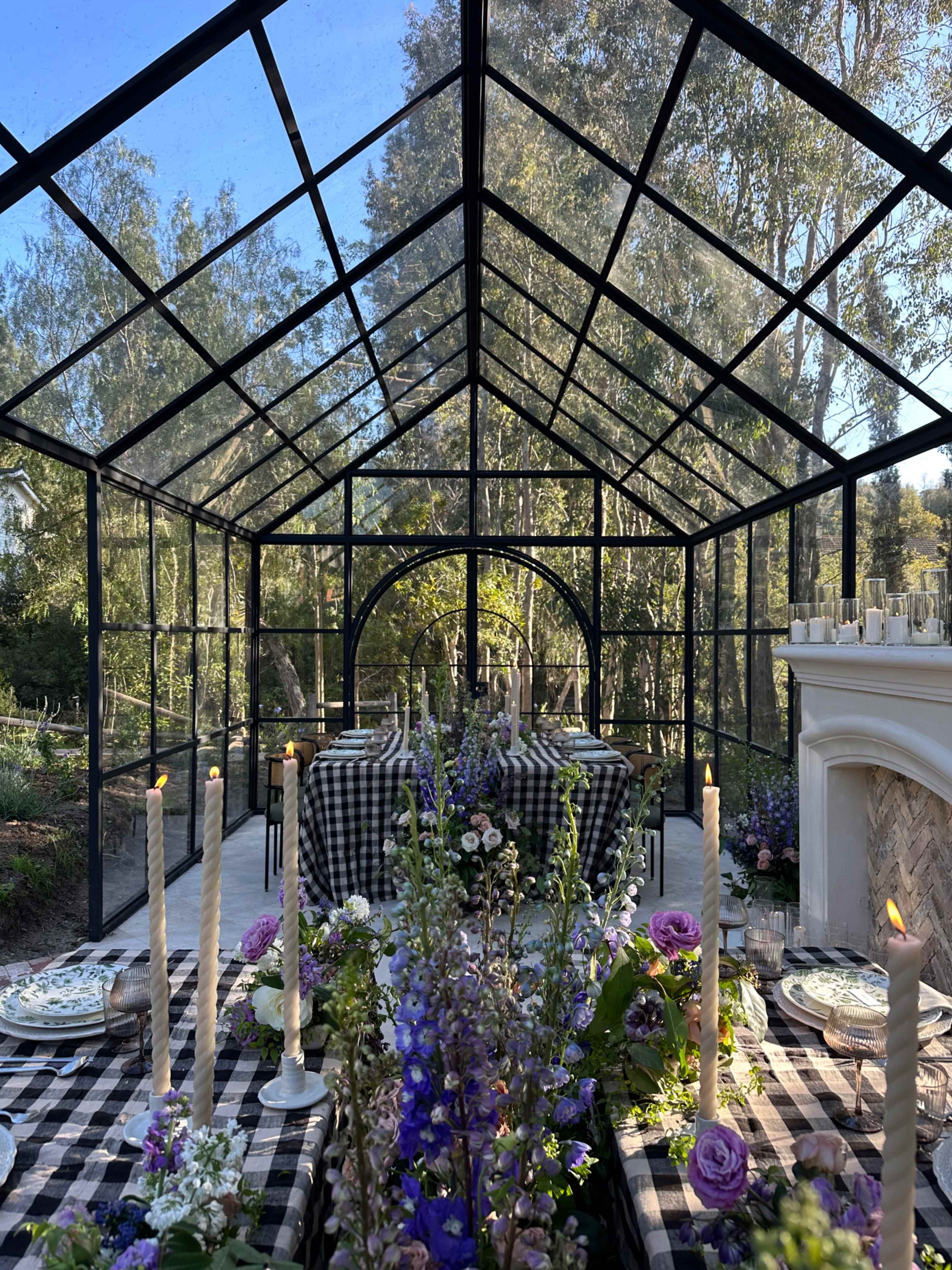 A glass greenhouse features a dining setup with checkered tablecloths, floral centerpieces, and candles, surrounded by greenery.