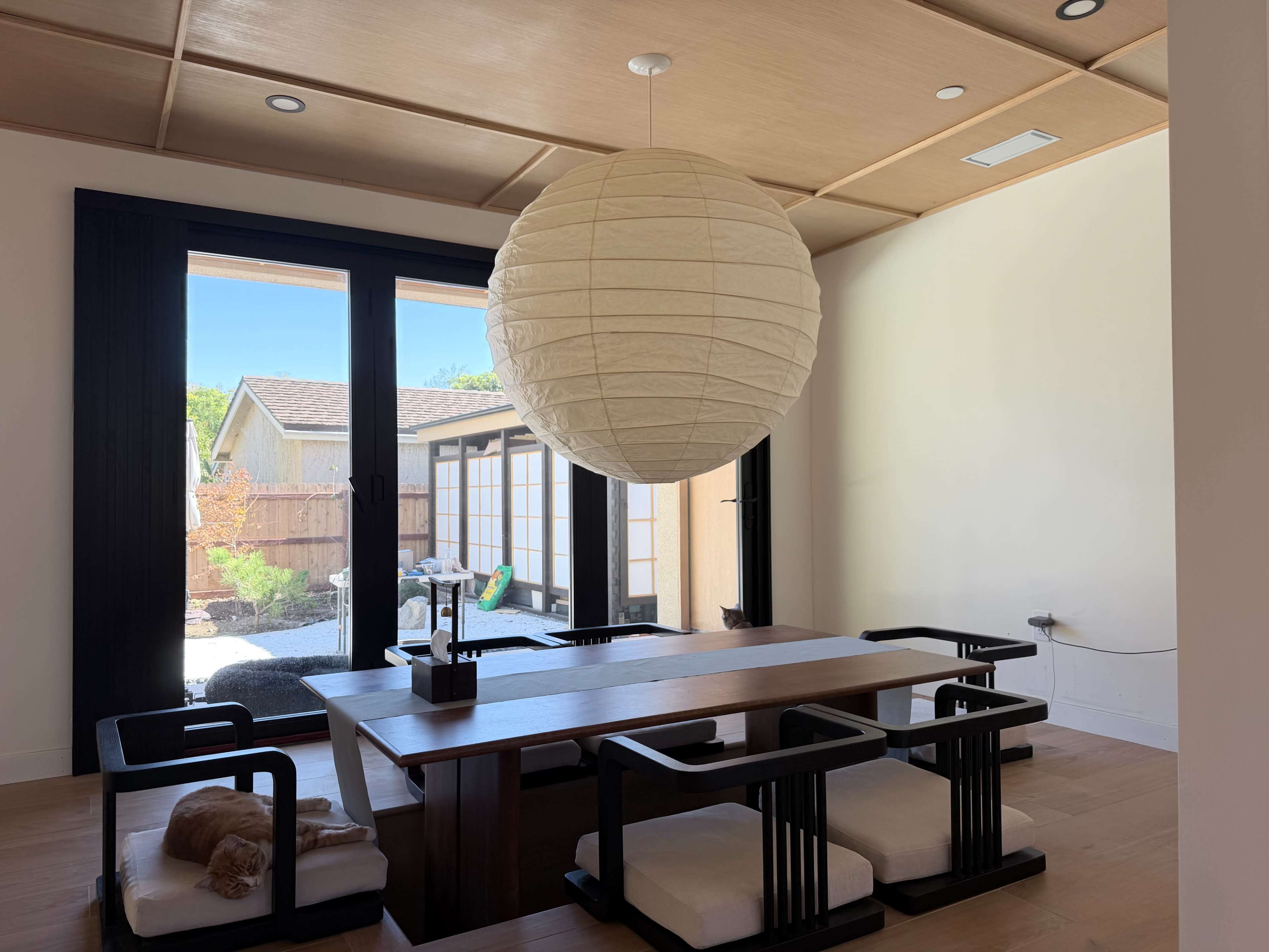 A modern dining room features a large paper lantern above a wooden table surrounded by black chairs, with sliding glass doors leading to a backyard.