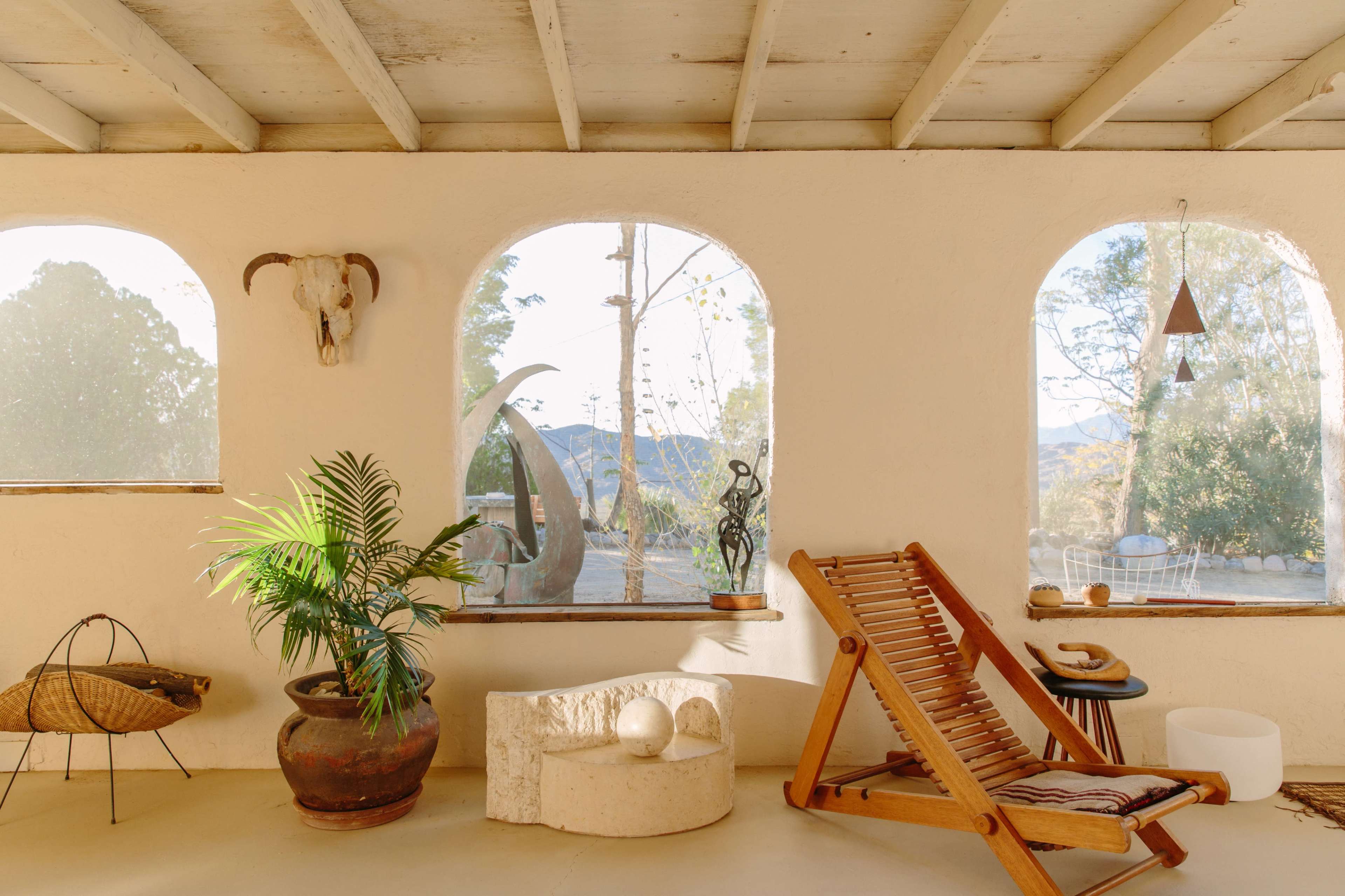 The image shows a spacious interior with arched windows, a wooden lounge chair, and various decorative elements, including a plant and sculptures, set against a backdrop of nature outside.