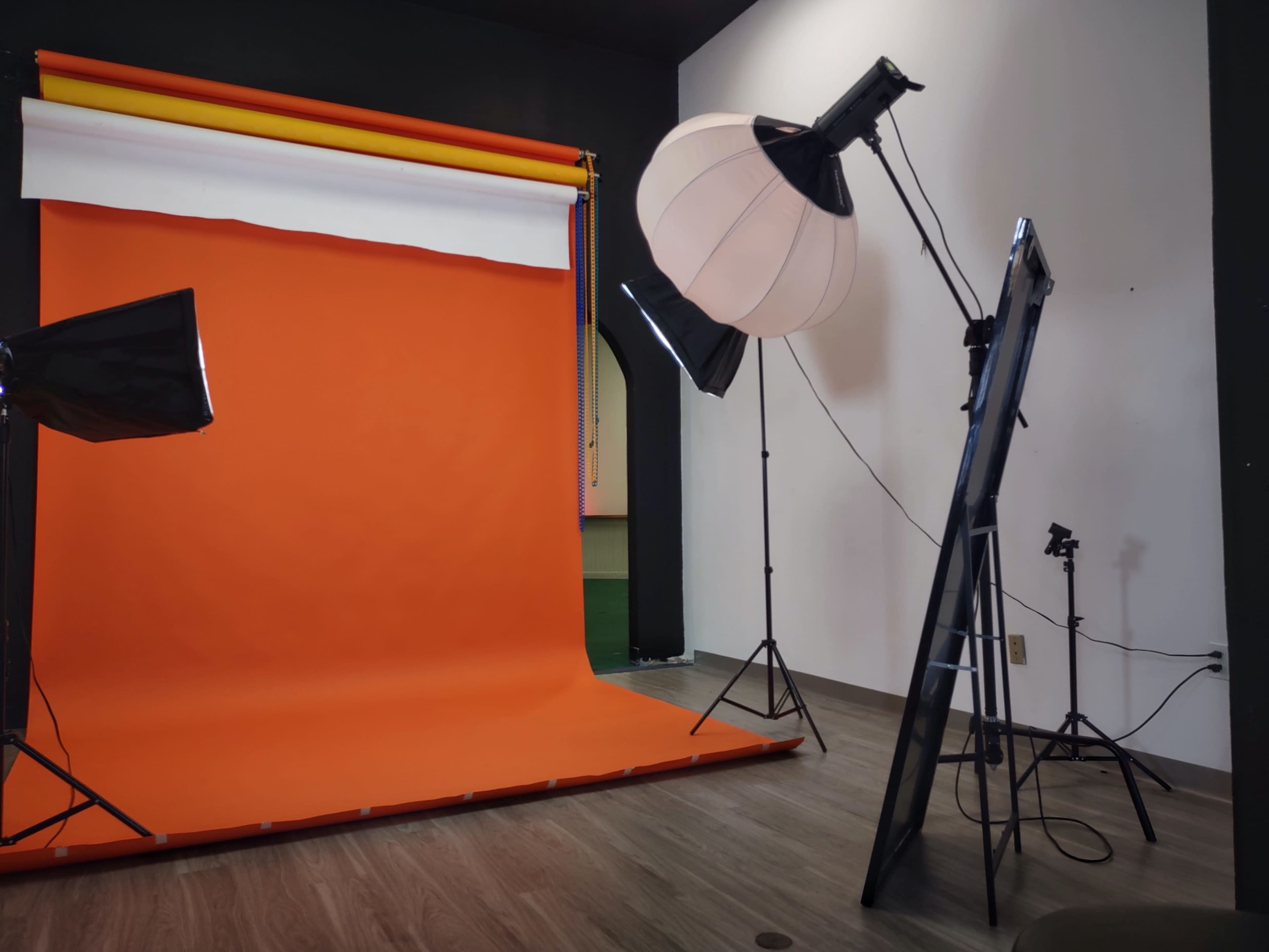 The image shows a photography studio with an orange backdrop, lighting equipment, and a large softbox set up for a photoshoot.