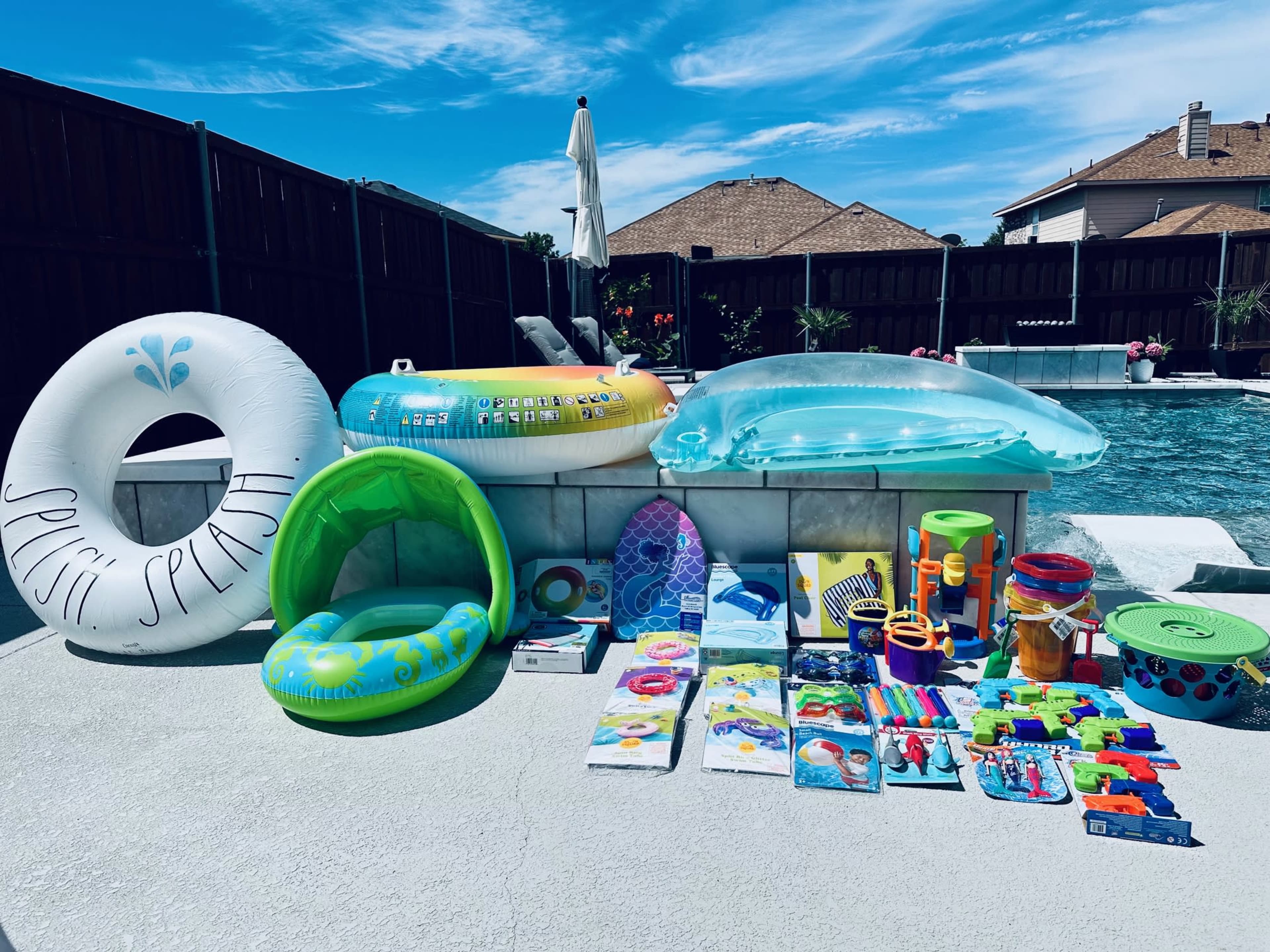 The image shows a variety of inflatable pool toys and floaties arranged next to a swimming pool on a sunny day.