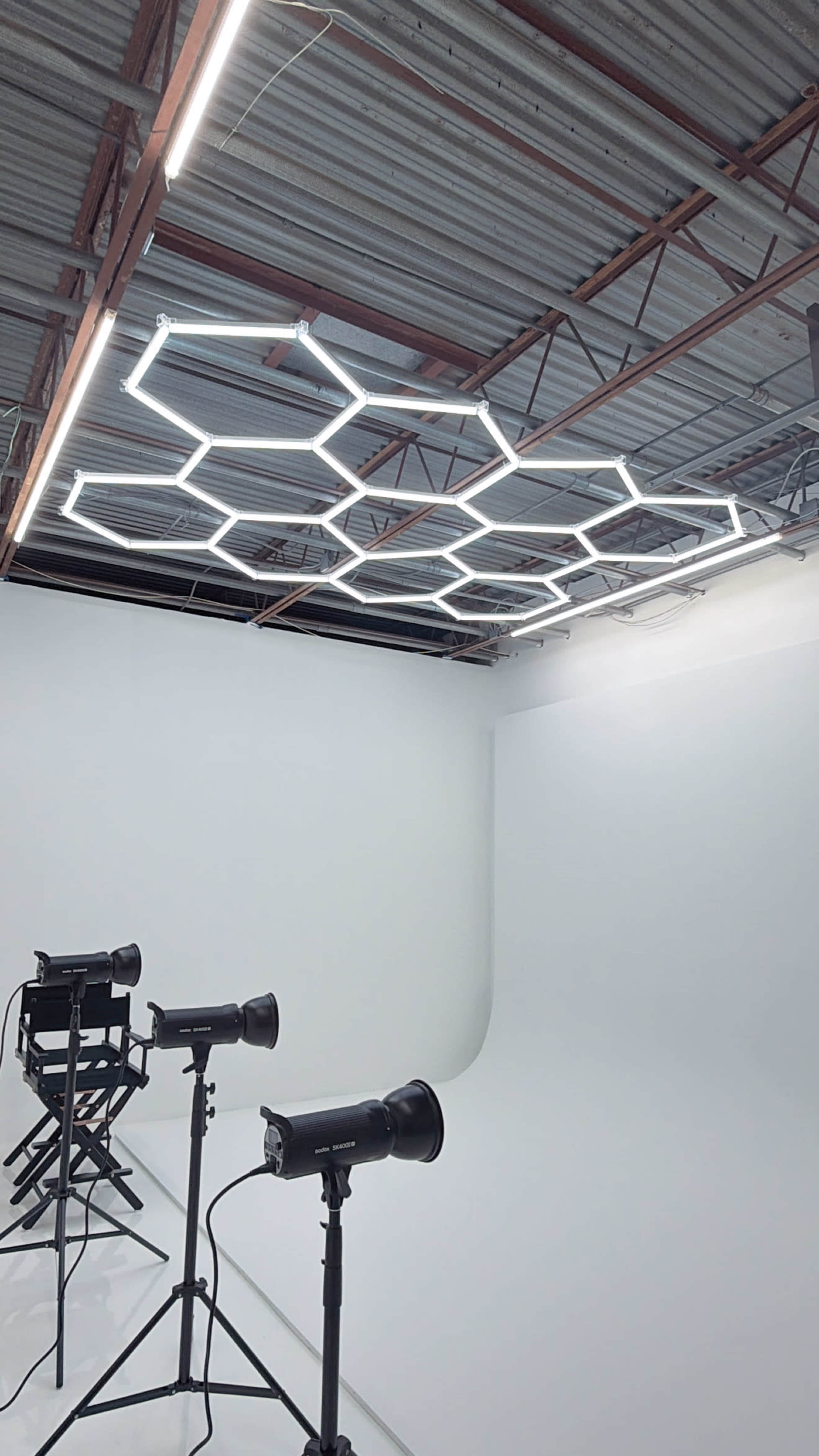 The image shows a minimalist photography studio with a hexagonal LED light fixture on the ceiling and two studio lights positioned on the floor.