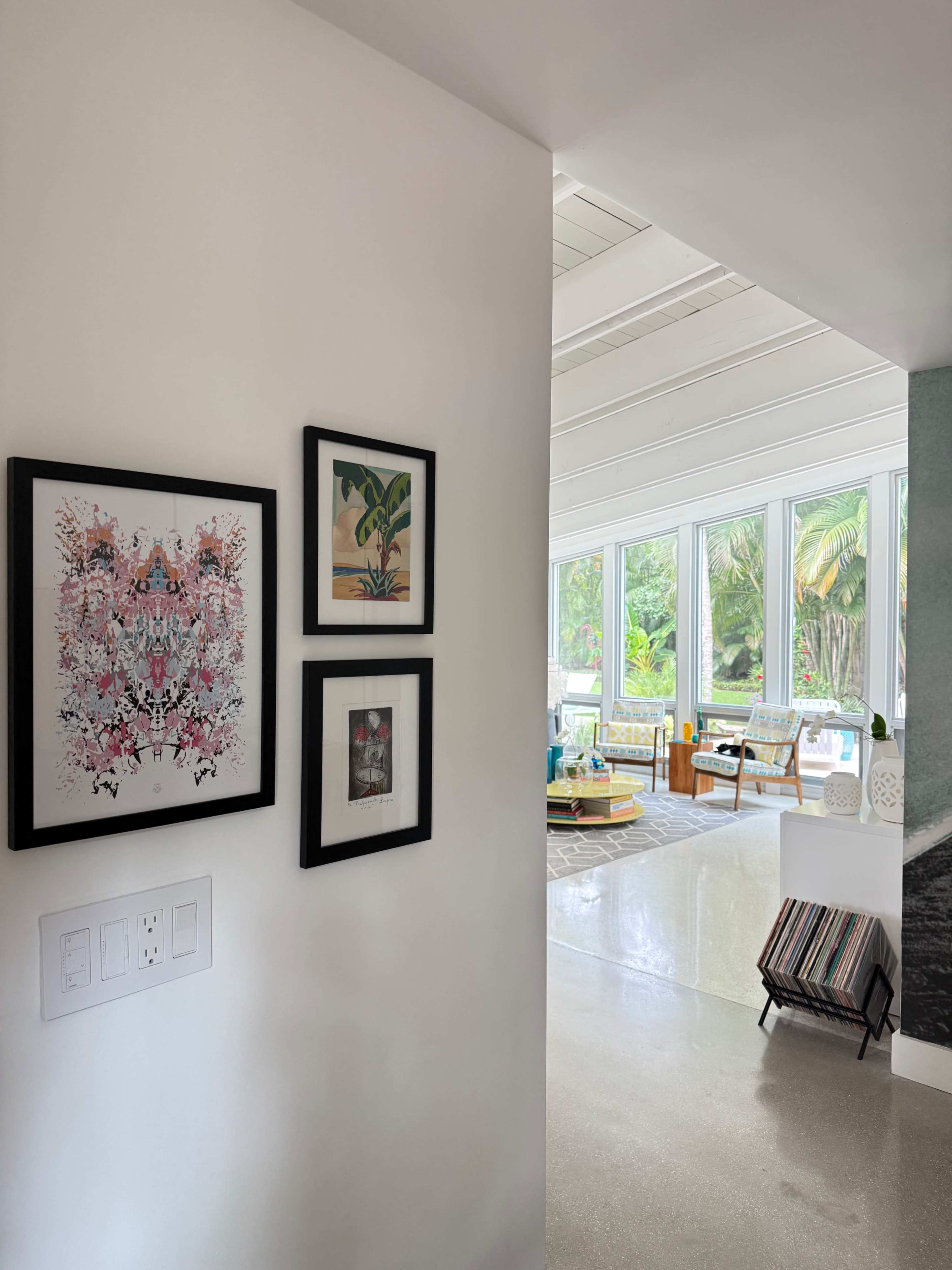 The image features a partially visible interior hallway leading to a bright room filled with large windows, showcasing greenery outside, and decorated with framed artwork on the wall.