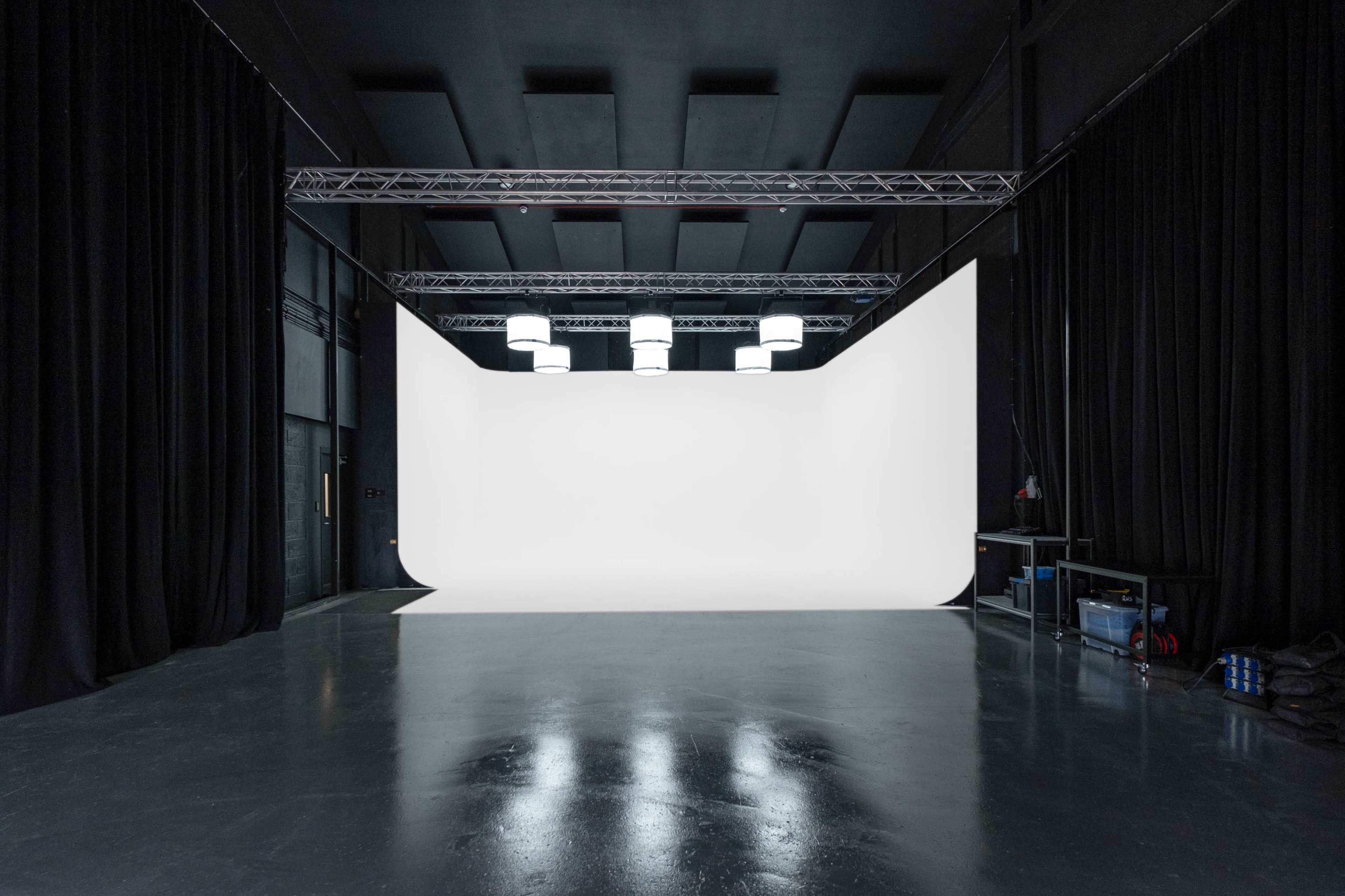 A spacious photography studio with a white backdrop, overhead lighting, and black curtains along the walls.
