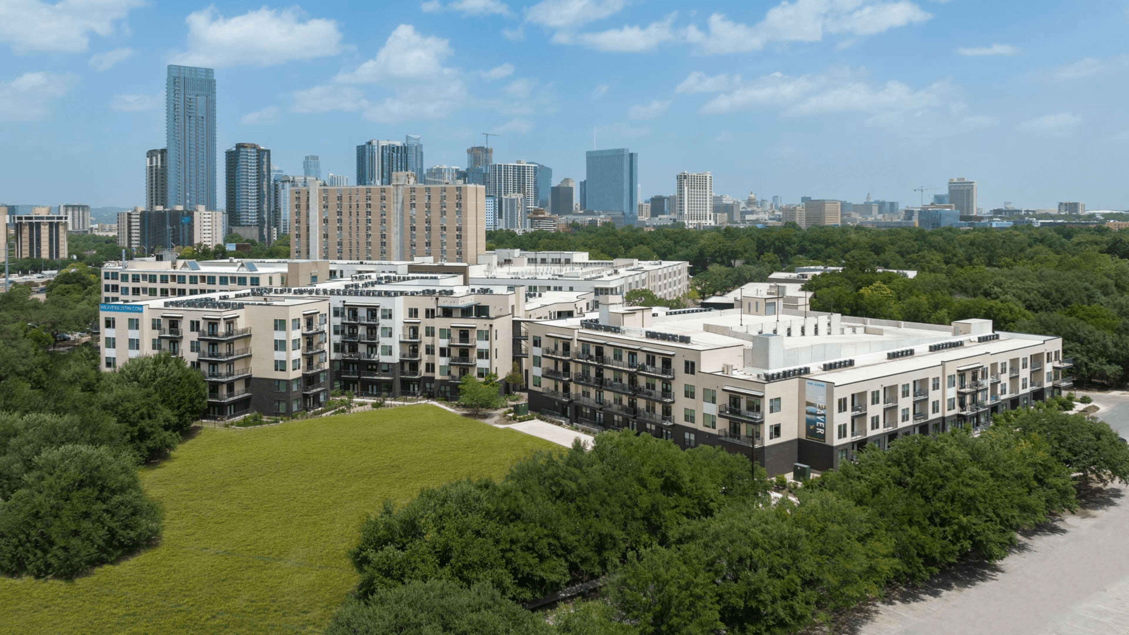 A suburban apartment complex surrounded by greenery, with a city skyline in the background.