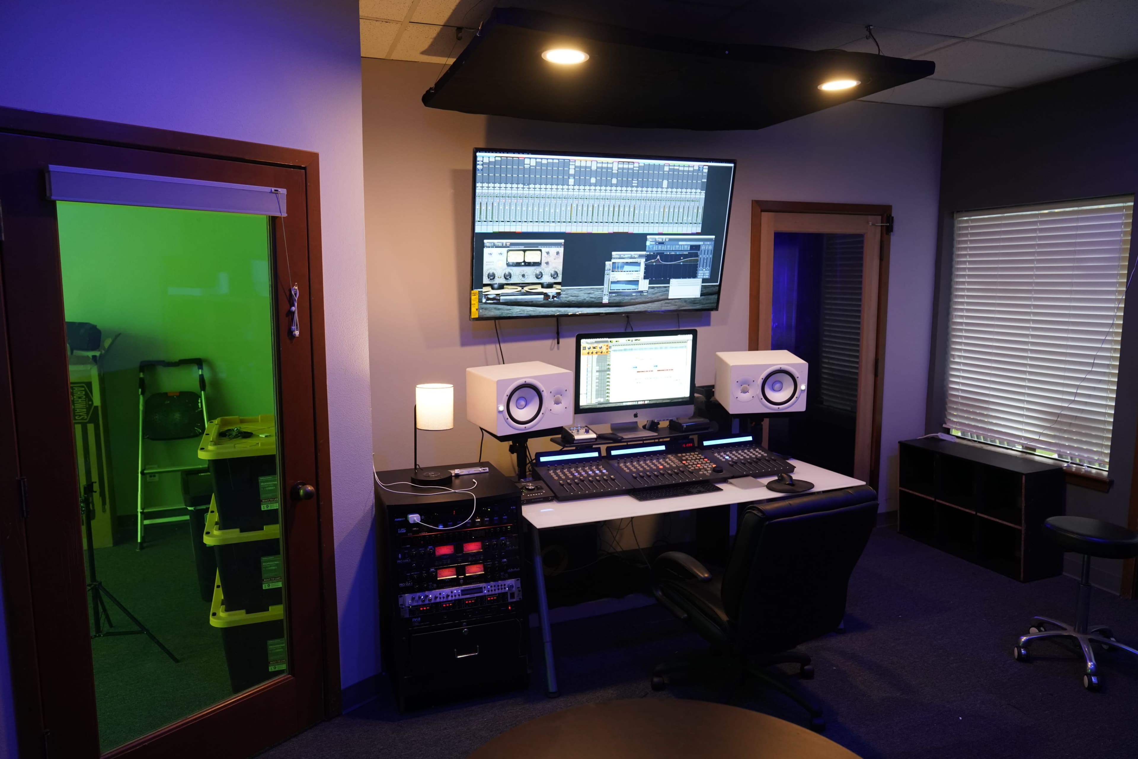 A modern recording studio features a desk with audio equipment, two monitors, and a green room visible through a glass door.