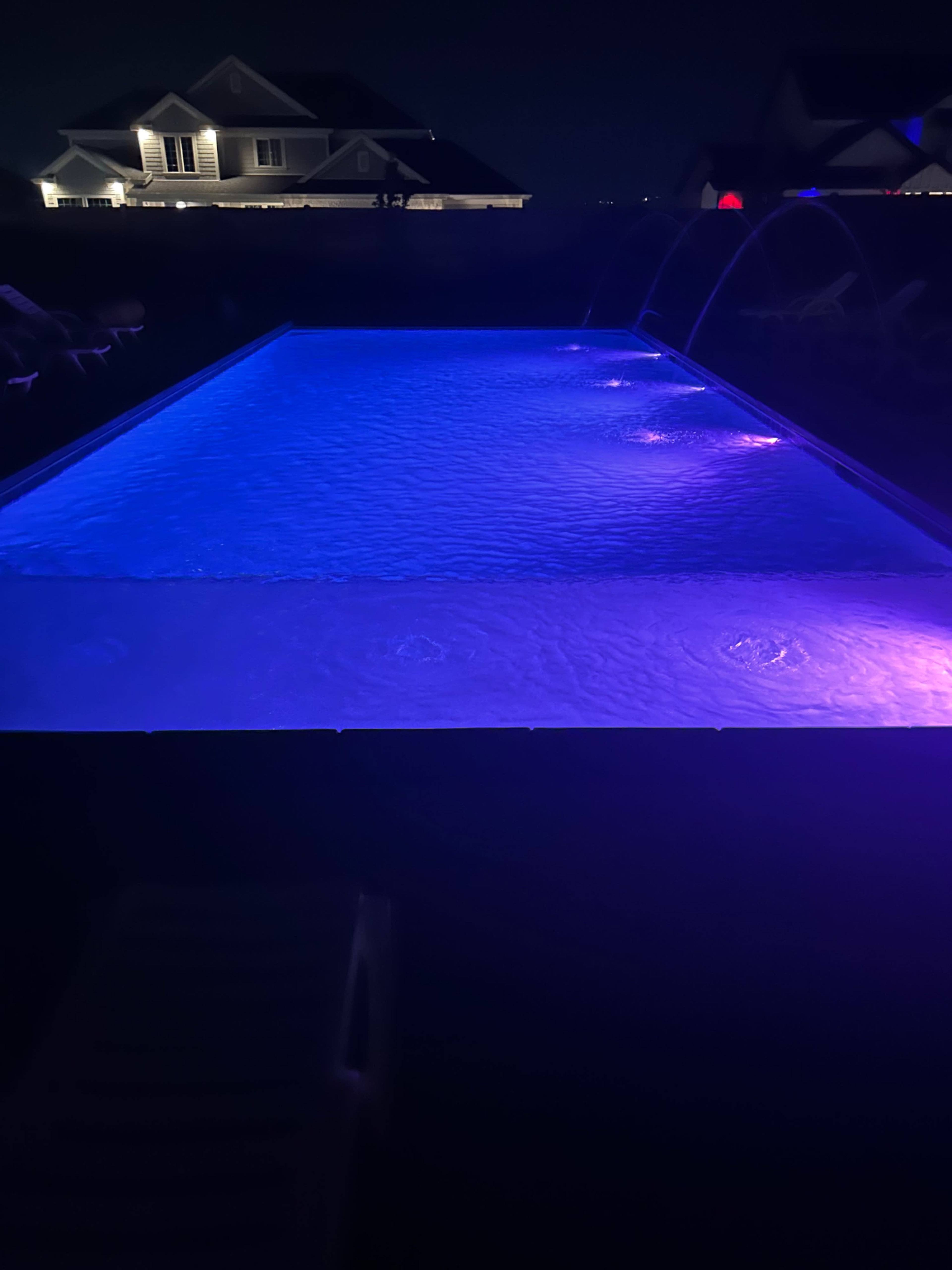A nighttime view of a swimming pool illuminated with blue and purple lights, surrounded by a residential area.