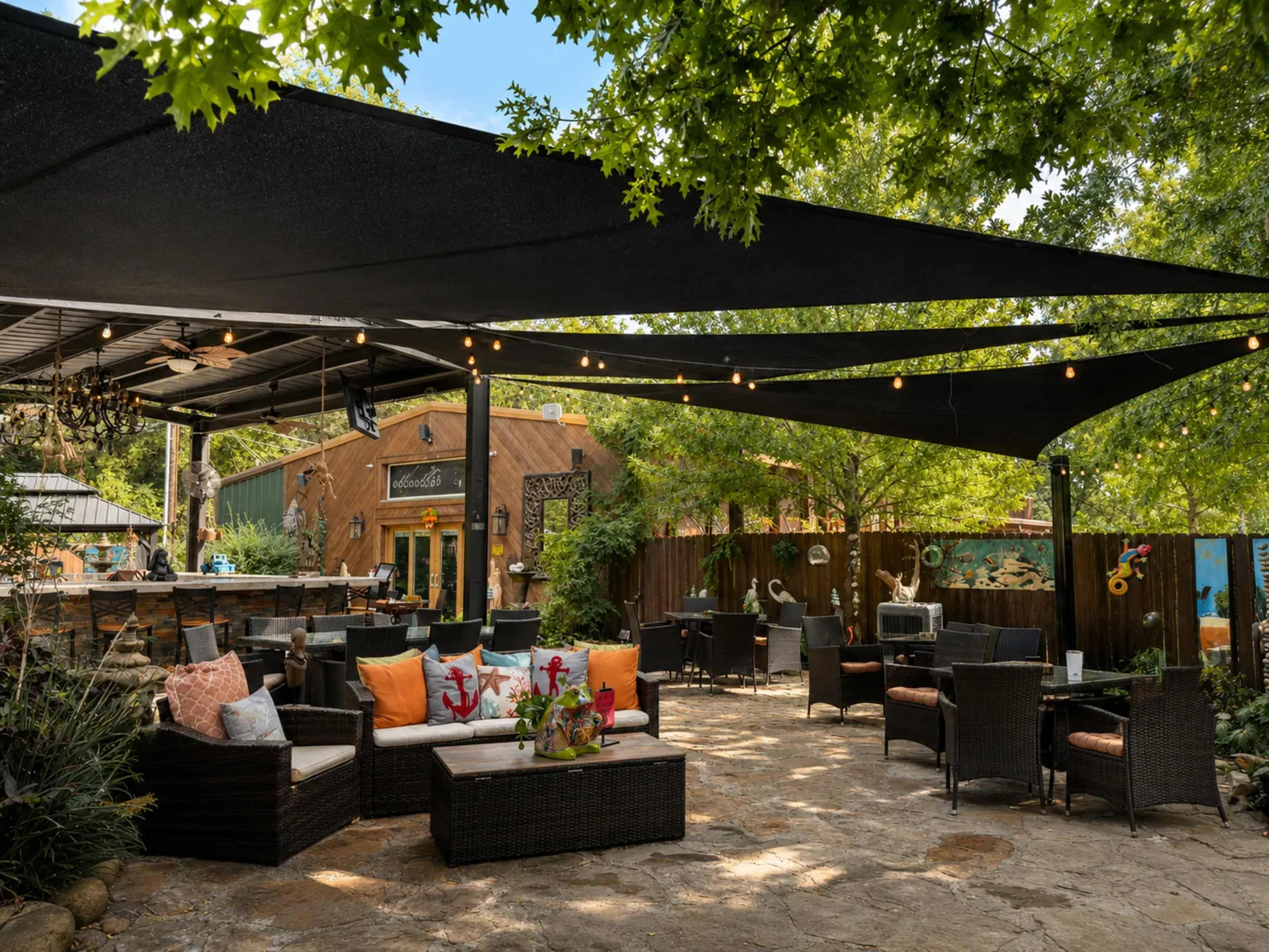The image shows a patio area with dark shade sails overhead, furnished with wicker seating and vibrant cushions amidst greenery and decorative elements.