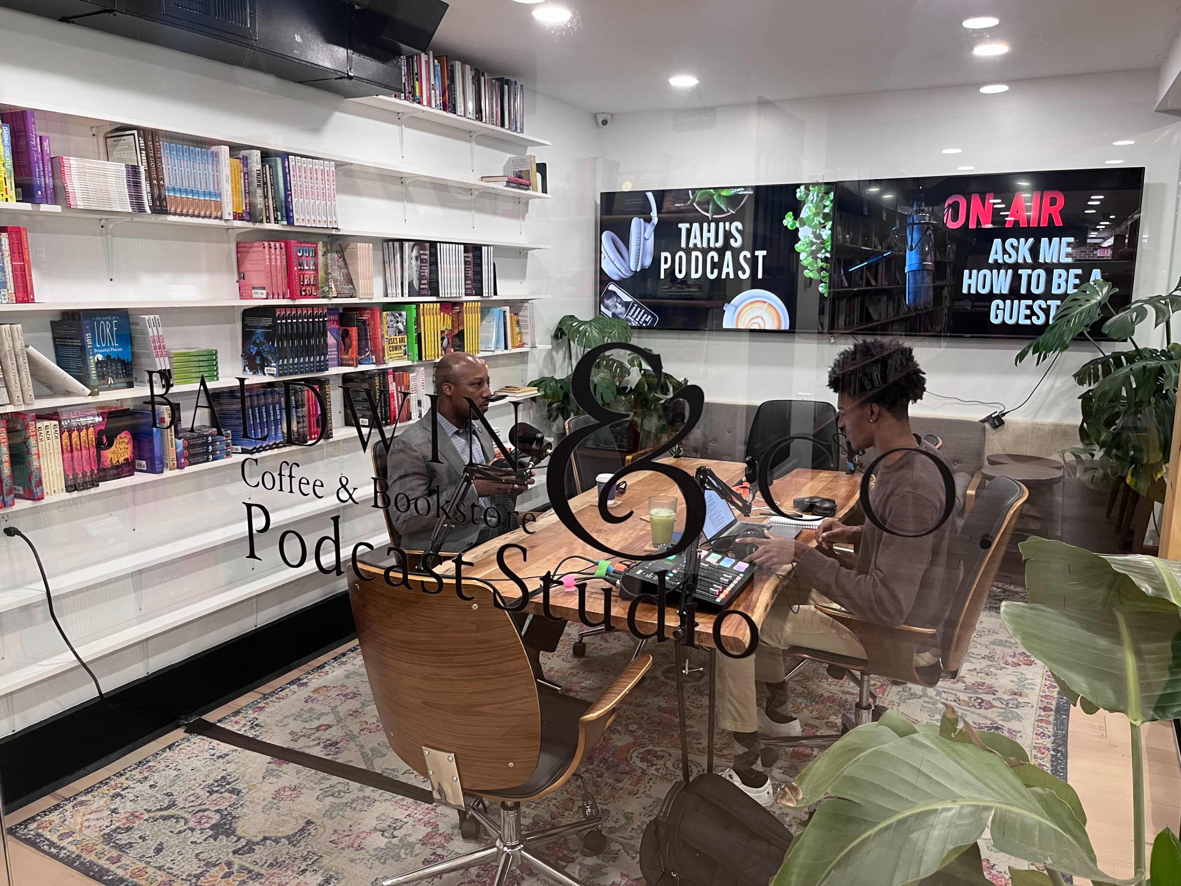 A man and a woman sit at a table in a podcast studio surrounded by books and a large screen displaying podcast information.