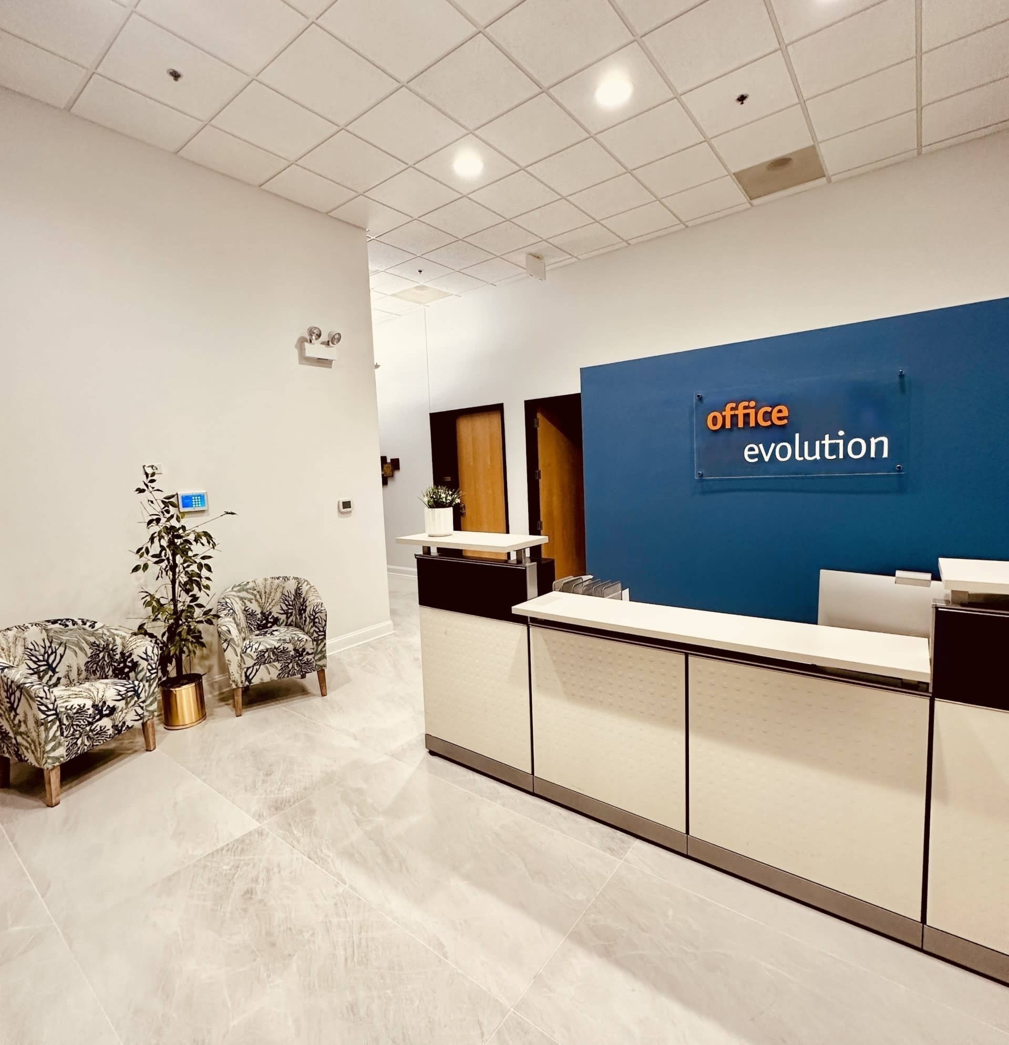 A modern reception area with a front desk, two patterned chairs, a potted plant, and an "office evolution" sign on the wall.