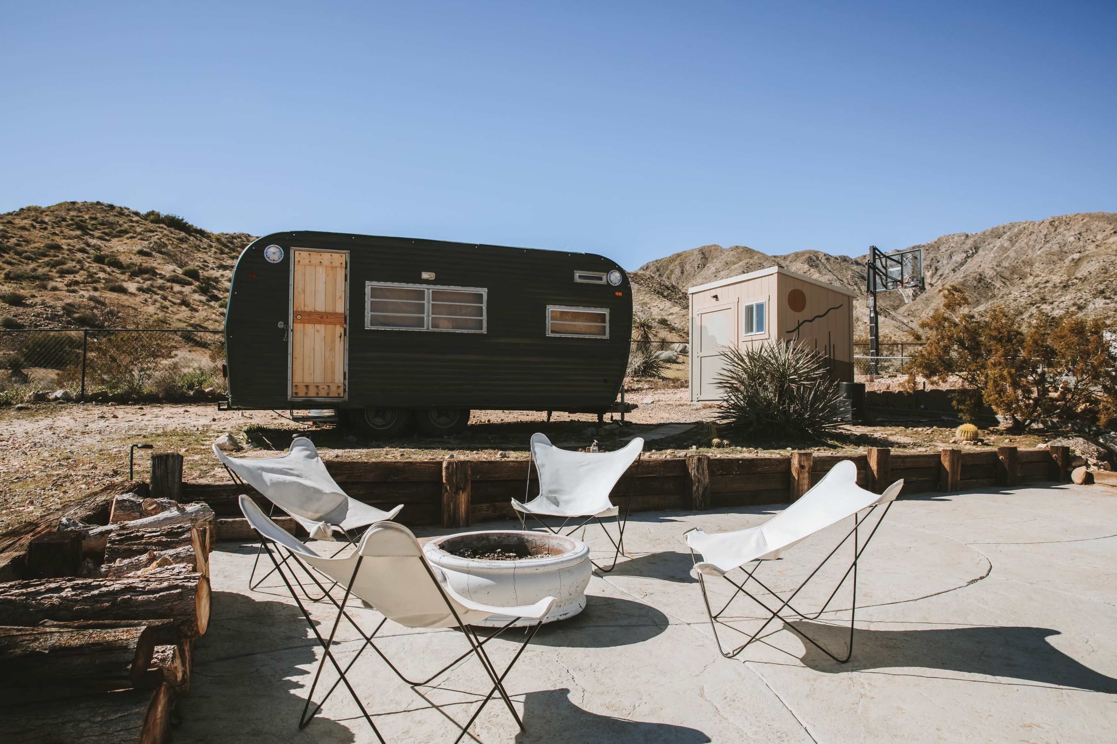 A vintage black trailer is parked beside a small structure in a desert landscape, surrounded by a fire pit and several butterfly chairs.