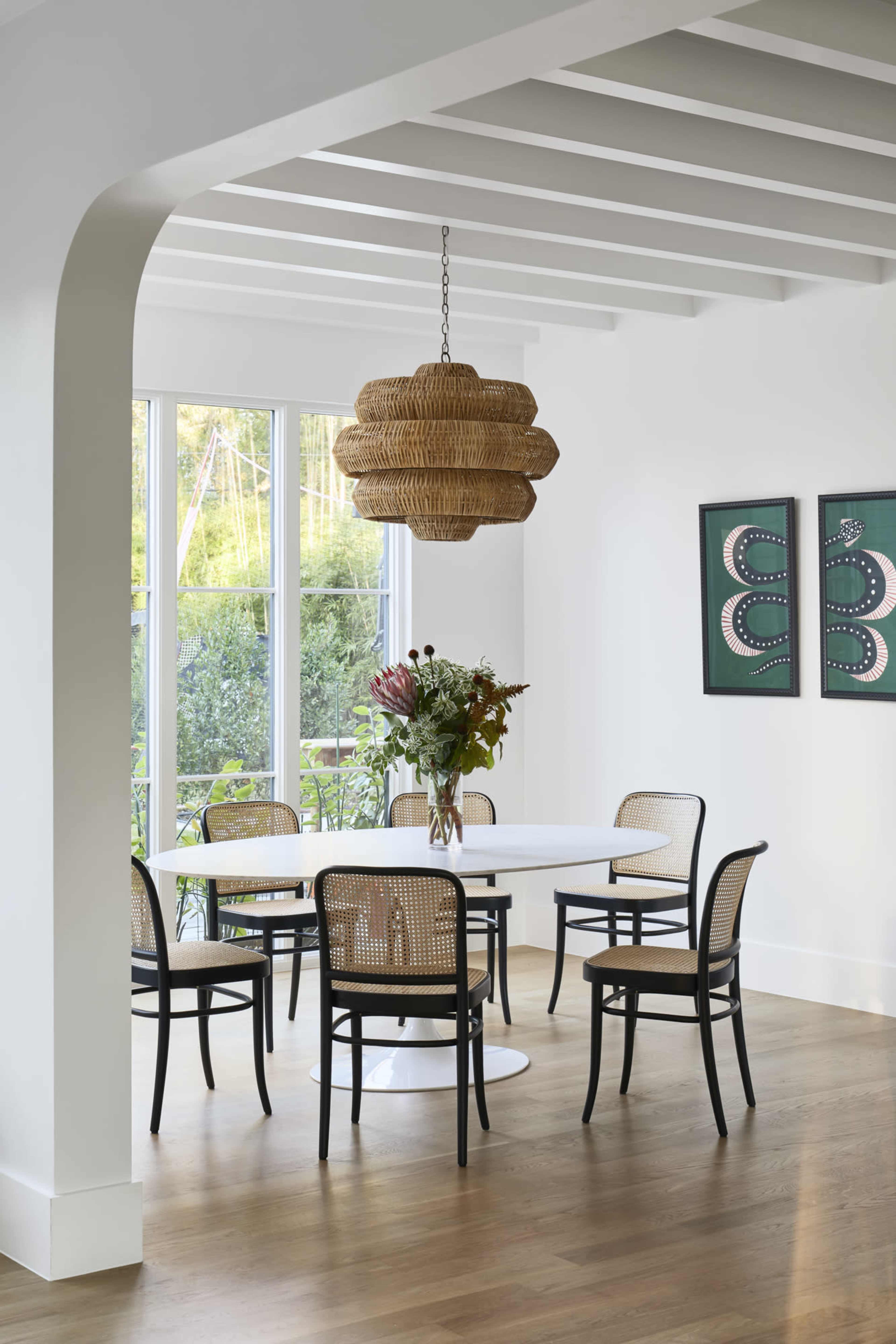 A modern dining room features a round white table surrounded by six black and cane chairs, with a woven pendant light hanging above.