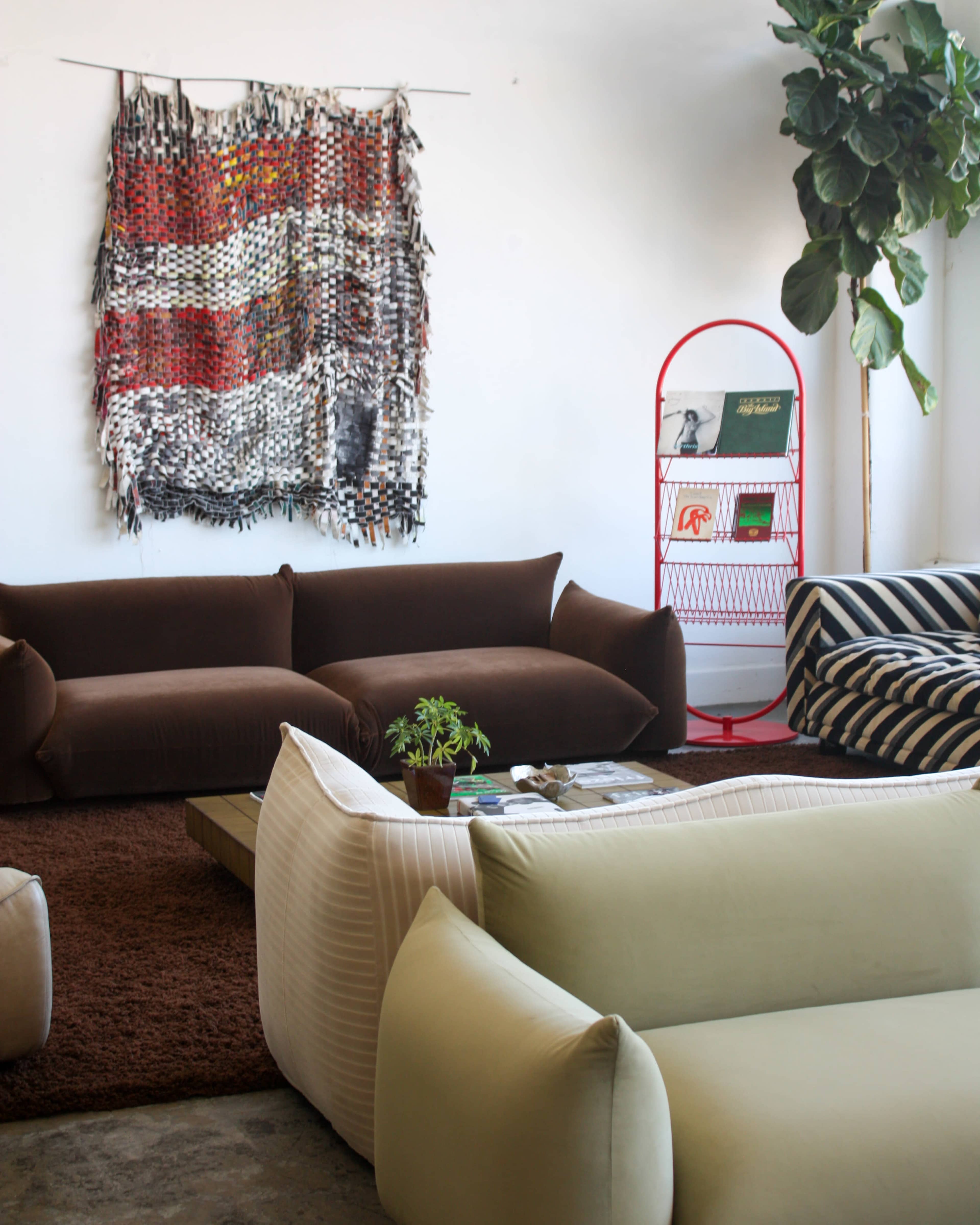The image features a modern living room with a mix of brown and green sofas, a large woven wall hanging, a red magazine rack, and a potted plant.