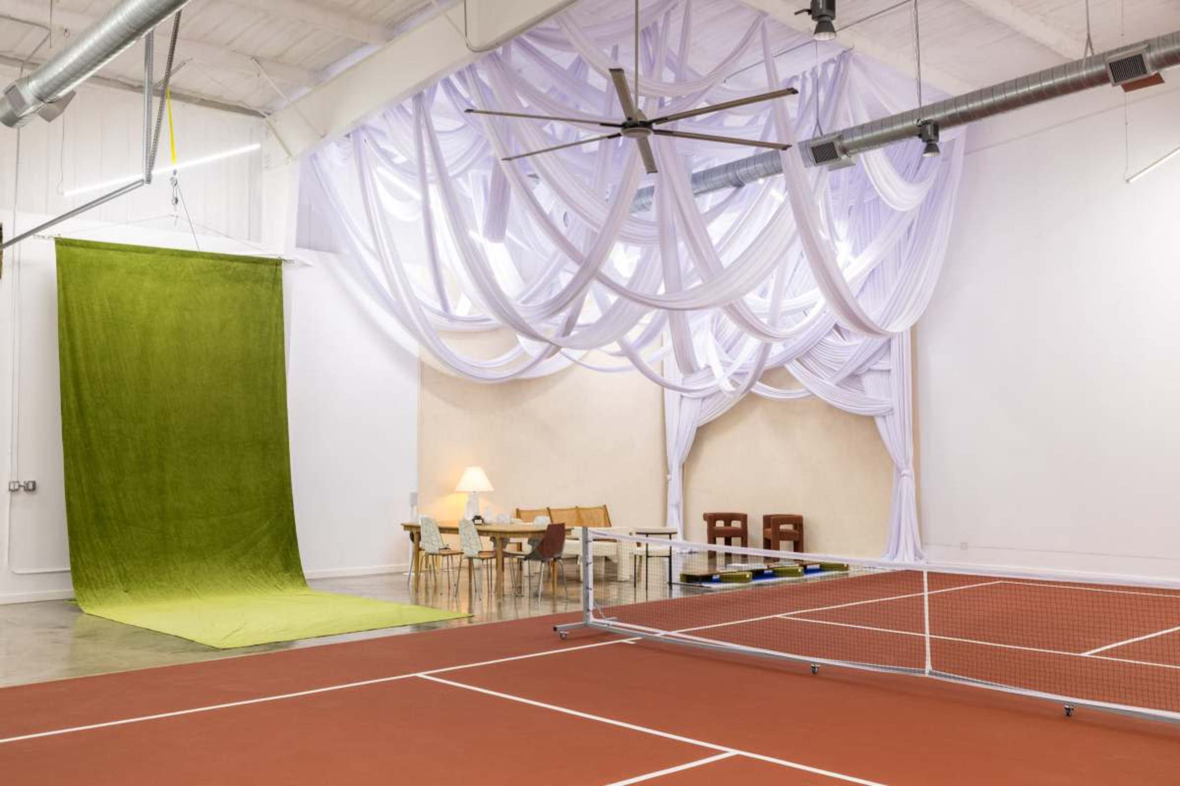 The image shows an indoor space with a red tennis court in the foreground, a green fabric draped on a wall, and a dining area featuring a table and chairs behind a curtain of white drapery.