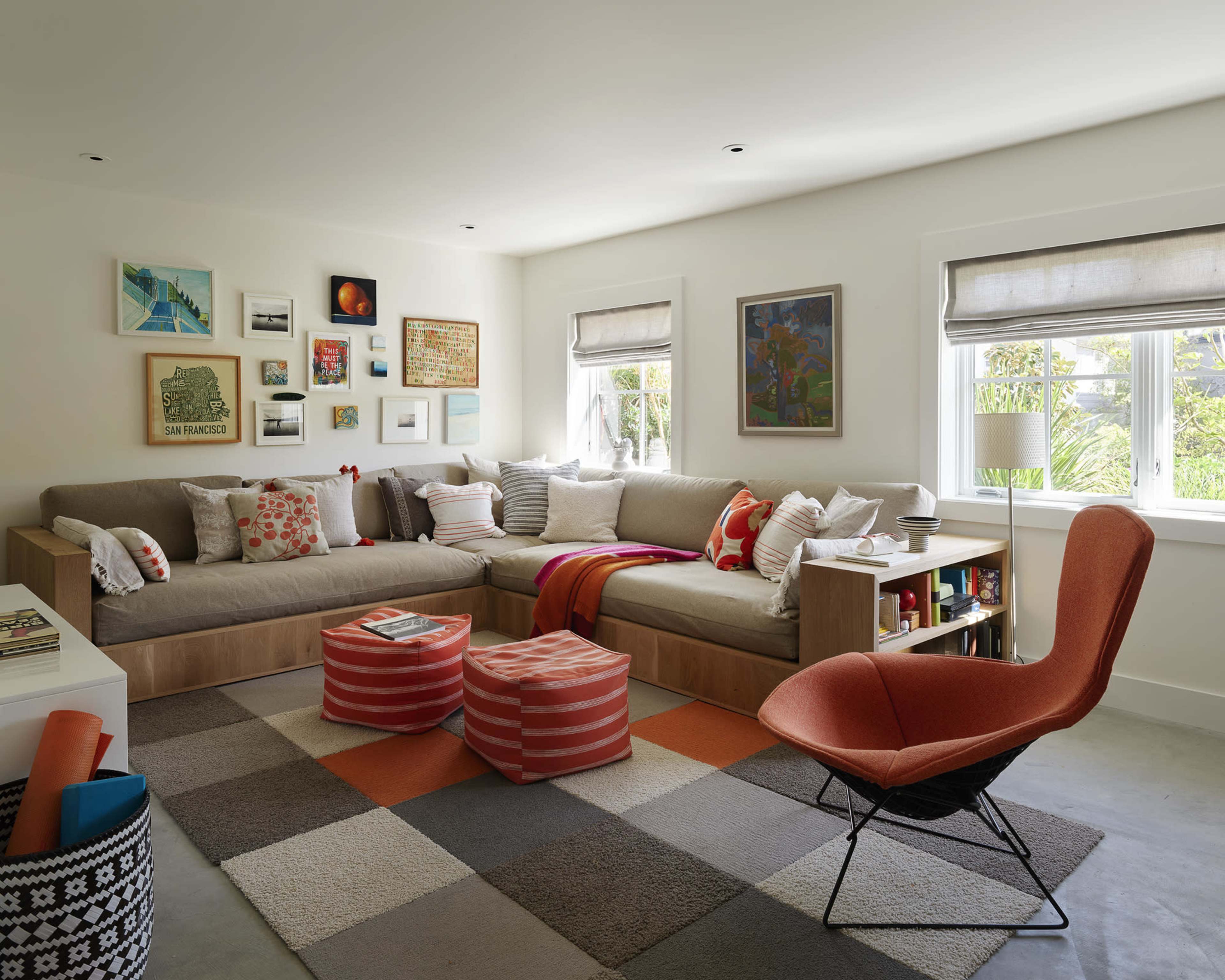 The image shows a modern living room with a sectional sofa, colorful cushions, a patterned rug, and a coffee table, illuminated by natural light through large windows.