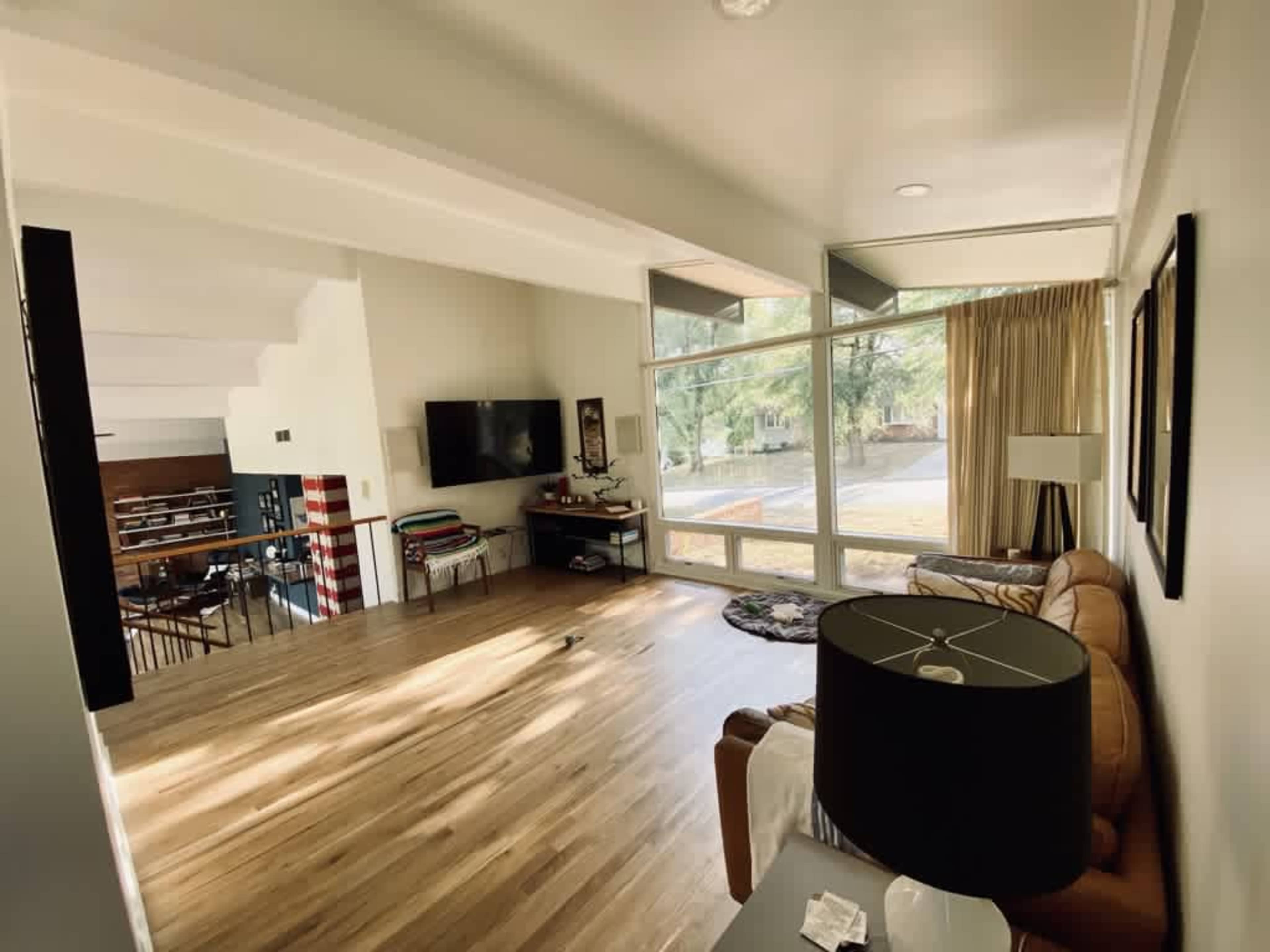 A modern living room features large windows, hardwood floors, and a sofa facing a wall-mounted television.