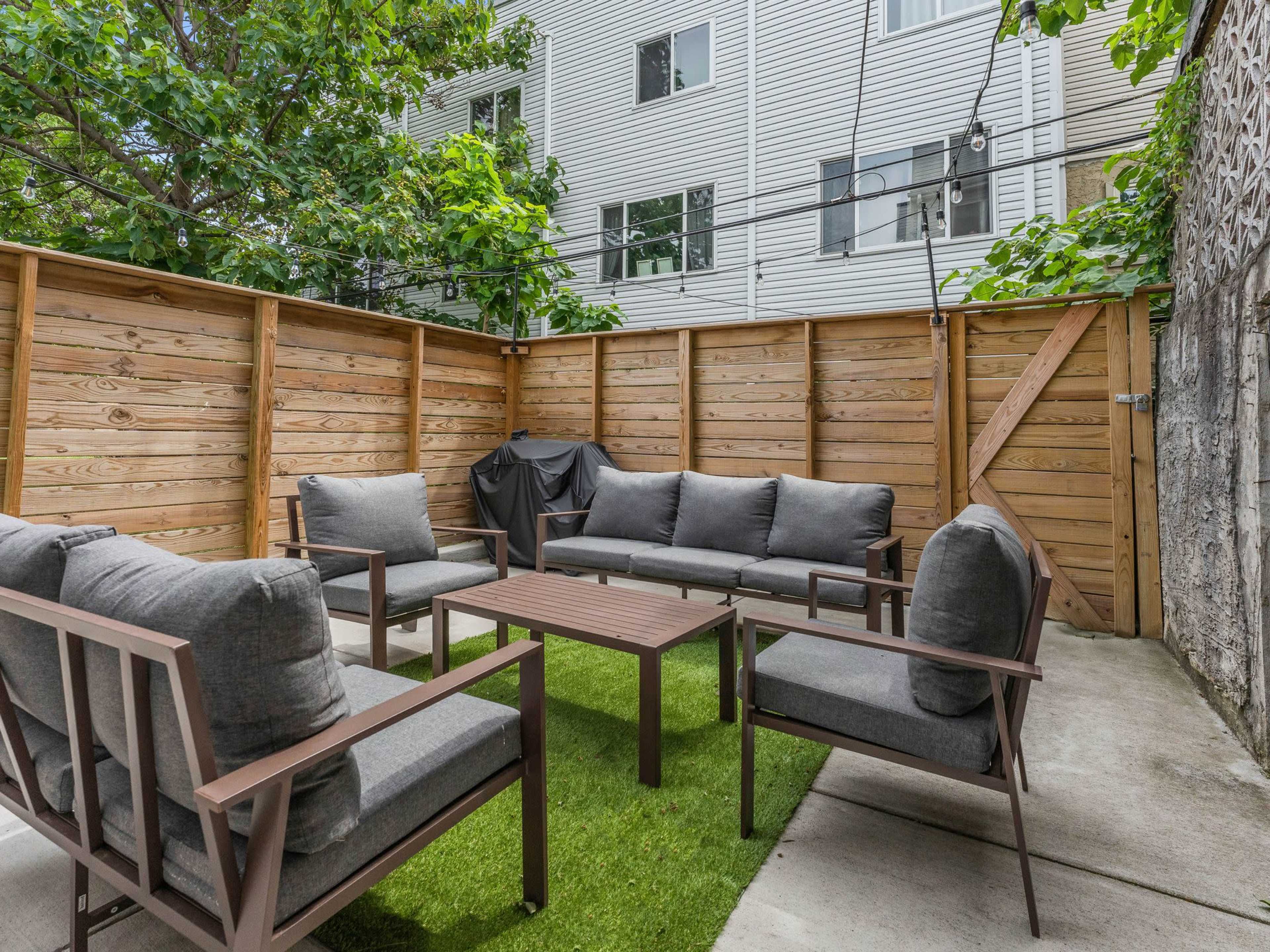 The image shows a seating area in a fenced outdoor space, featuring six gray cushioned chairs surrounding a wooden table on green grass.