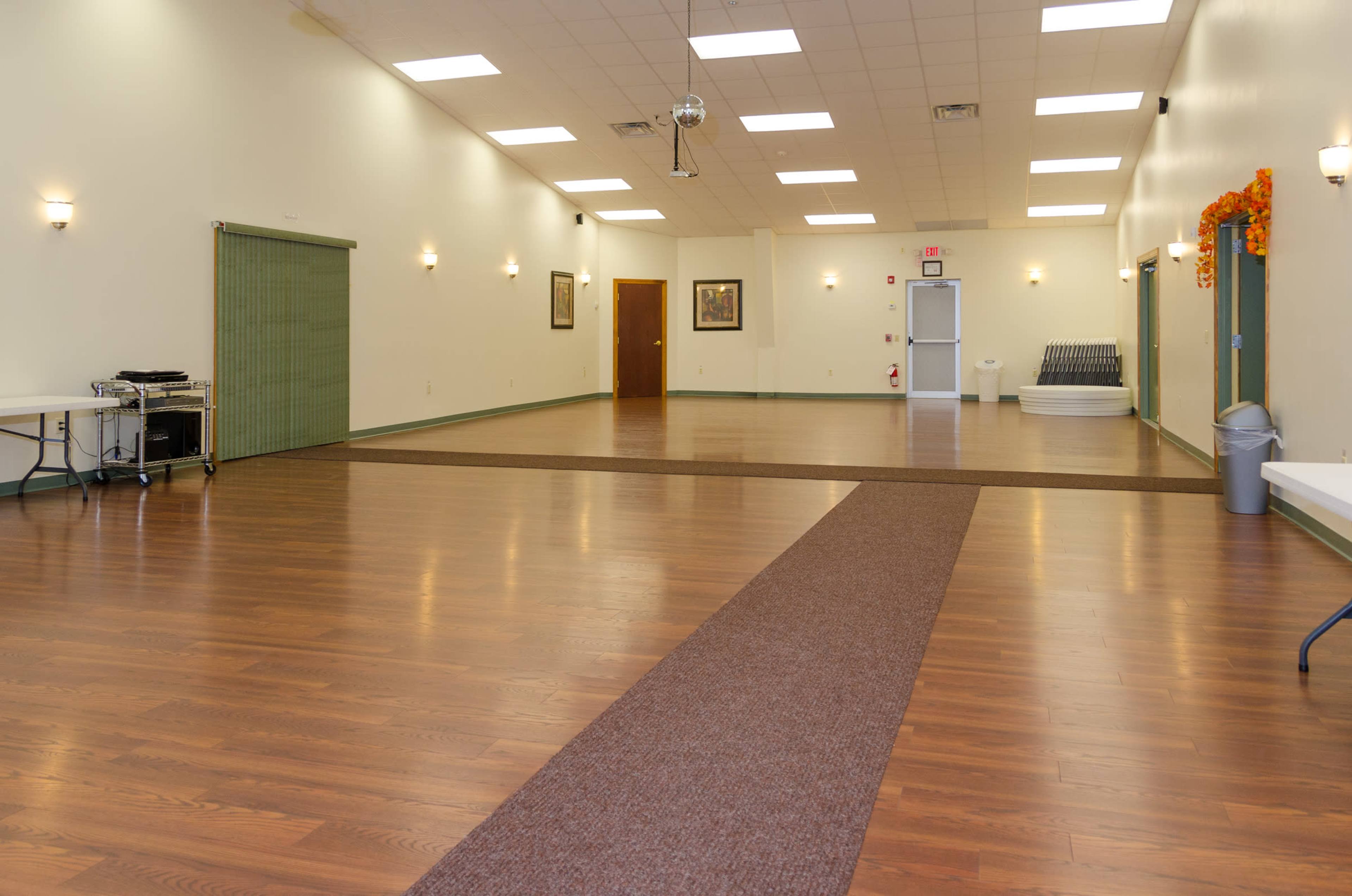 A spacious indoor venue with wooden flooring, green walls, and minimal furnishings, featuring a central carpeted walkway.