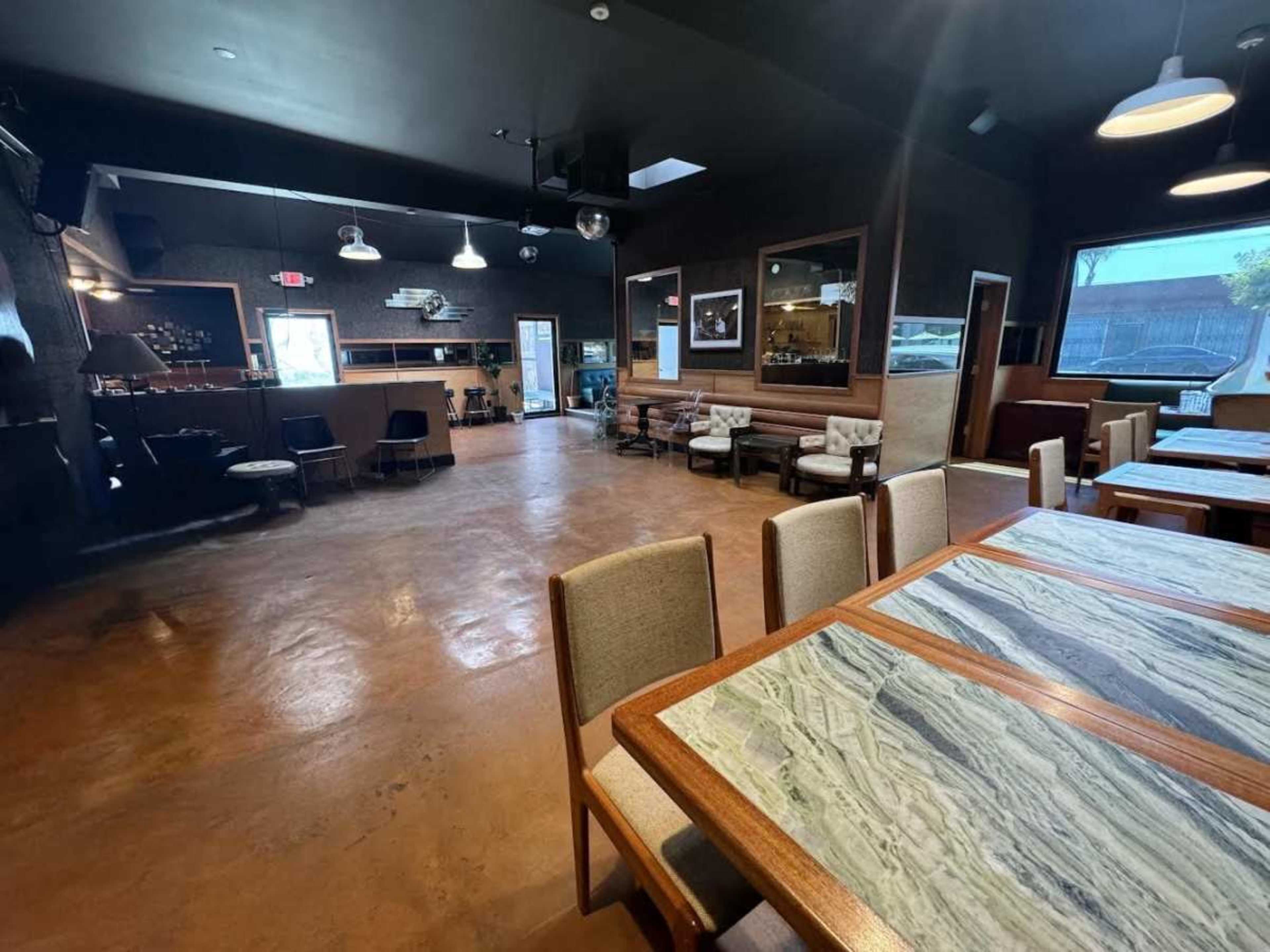 The image shows a spacious, modern interior of a restaurant or café with tables and chairs, a bar area in the background, and large windows allowing natural light.