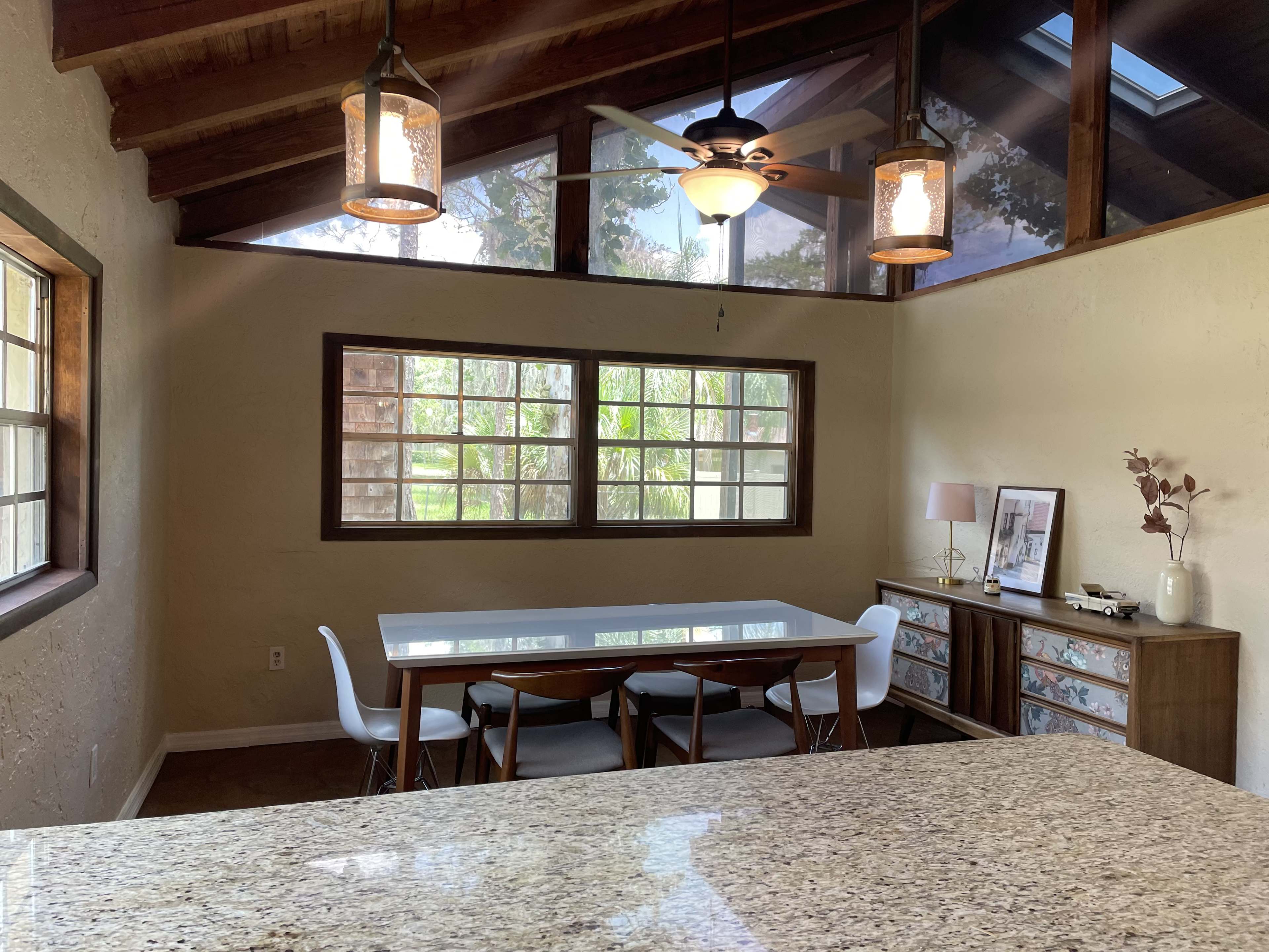 A dining area with a rectangular table surrounded by chairs, situated under a vaulted ceiling with large windows allowing natural light to enter.