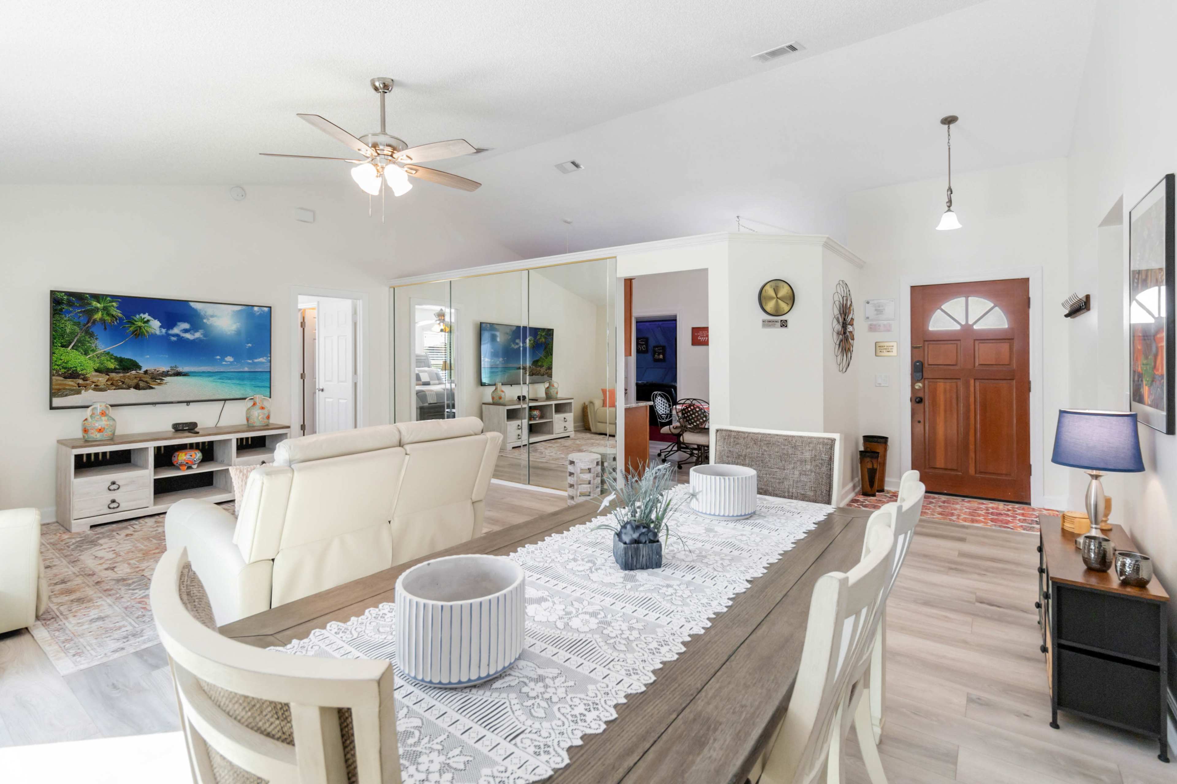 The image shows a light-filled living and dining area with a large television, comfortable seating, and a decorative dining table set with centerpieces.