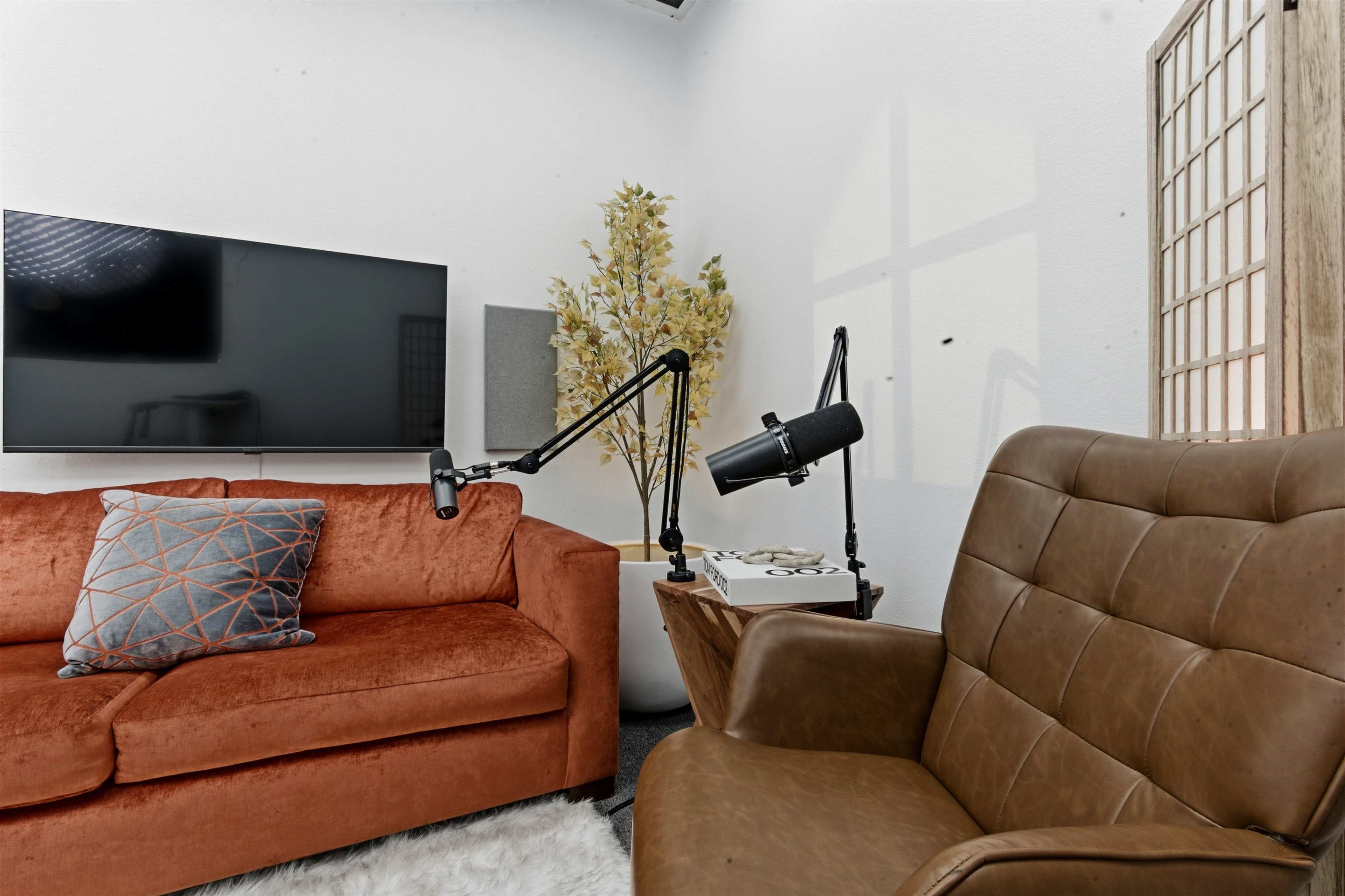 The image shows a cozy living room setup with an orange sofa, a brown armchair, a TV mounted on the wall, and a potted plant in the corner.