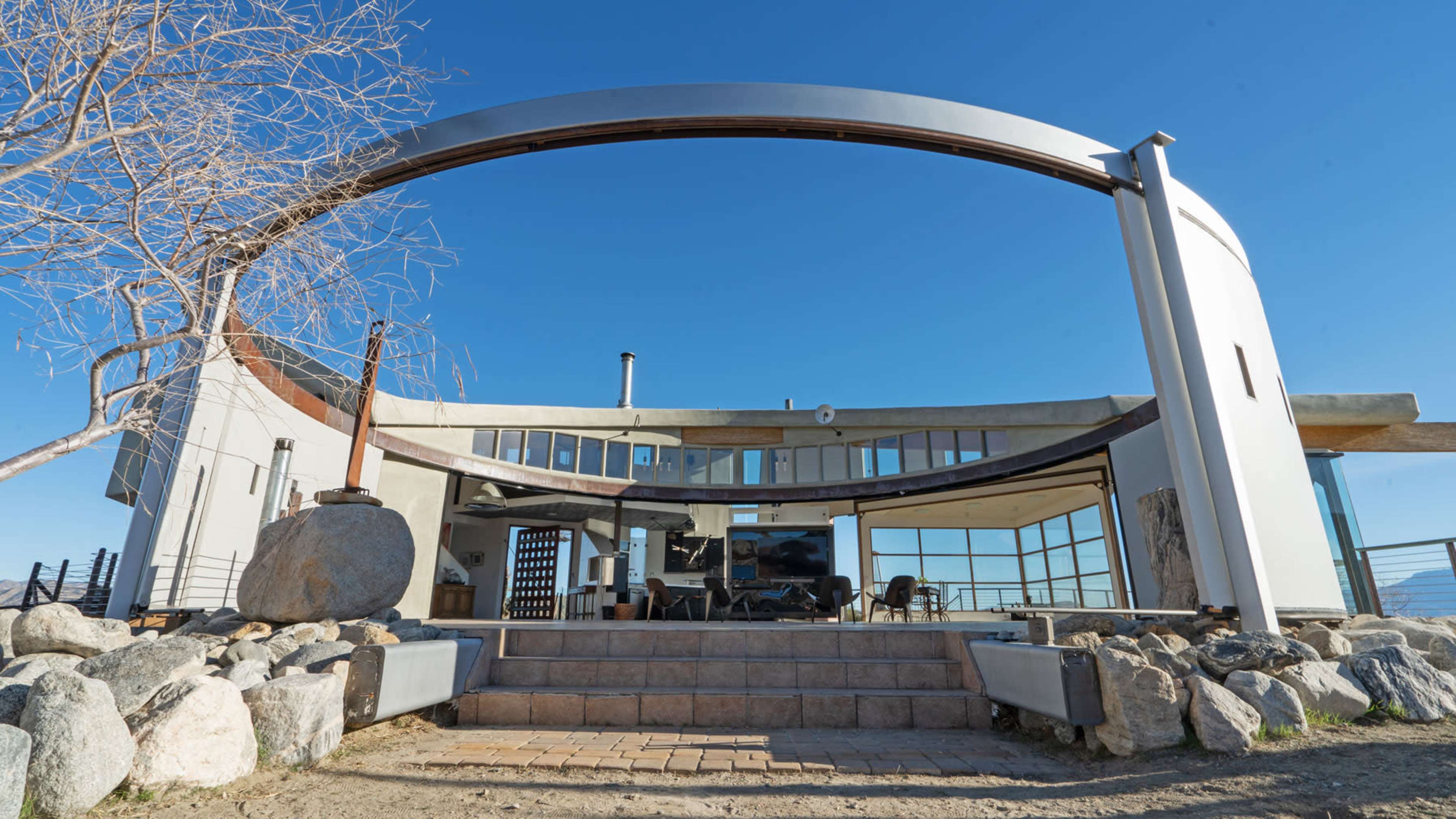 A modern, circular architectural design stands prominently against a clear blue sky, featuring expansive glass panels and a stone pathway leading to its entrance.