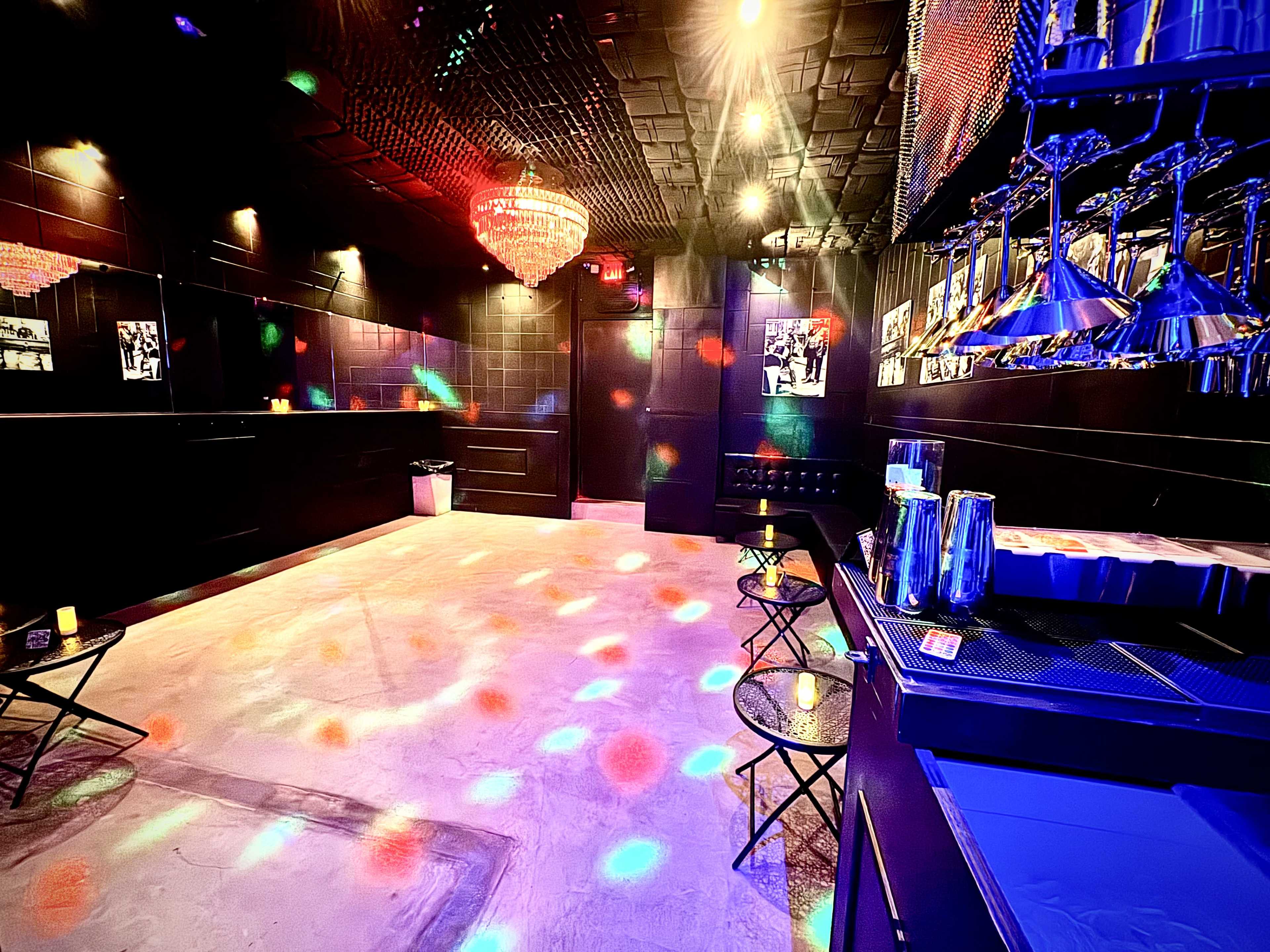 The image depicts a dimly lit lounge or bar area with colorful disco lights illuminating the floor and walls, and a bar featuring metallic fixtures and stools.