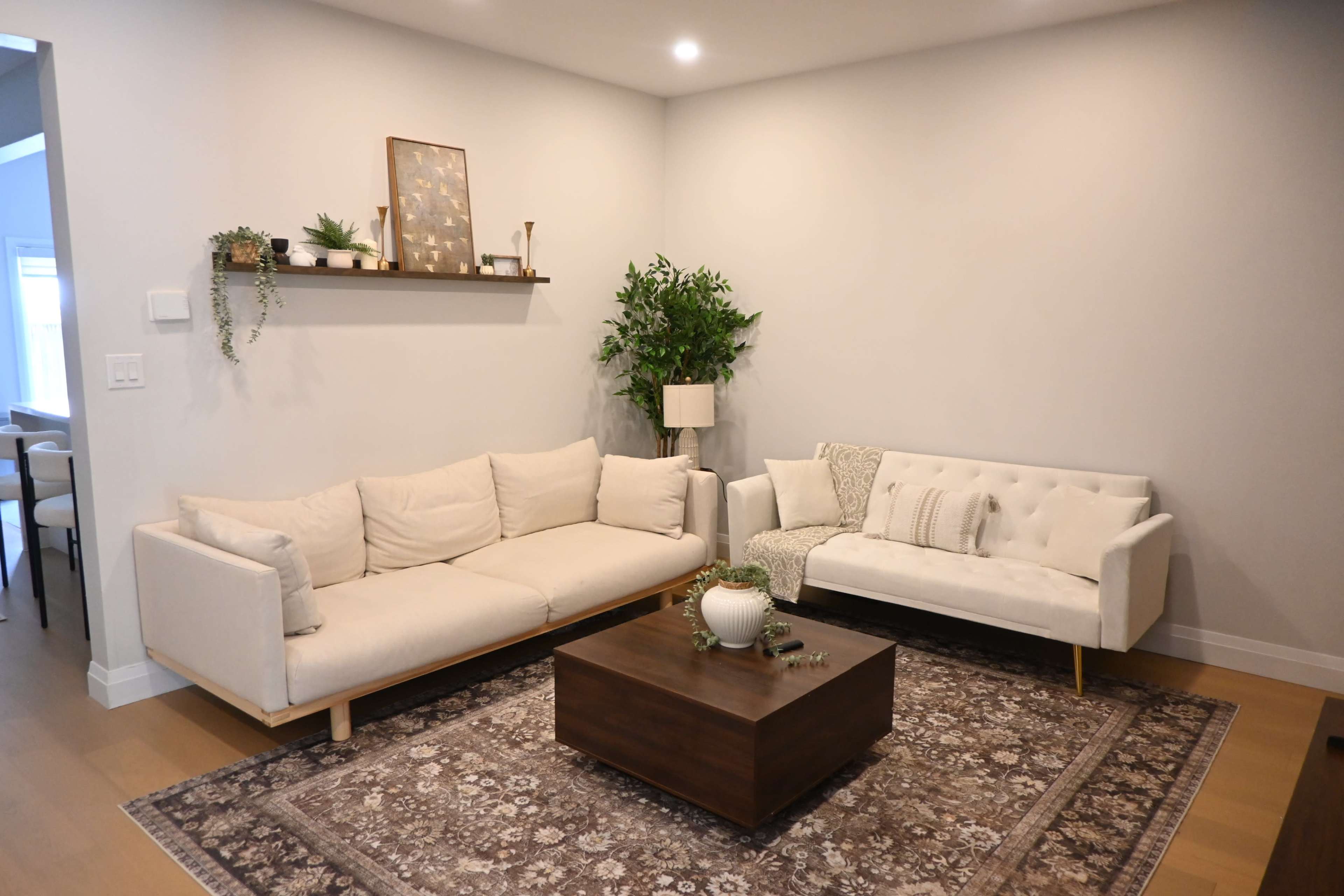 The image shows a modern living room with two beige sofas, a wooden coffee table on a decorative rug, and a potted plant against a light gray wall.