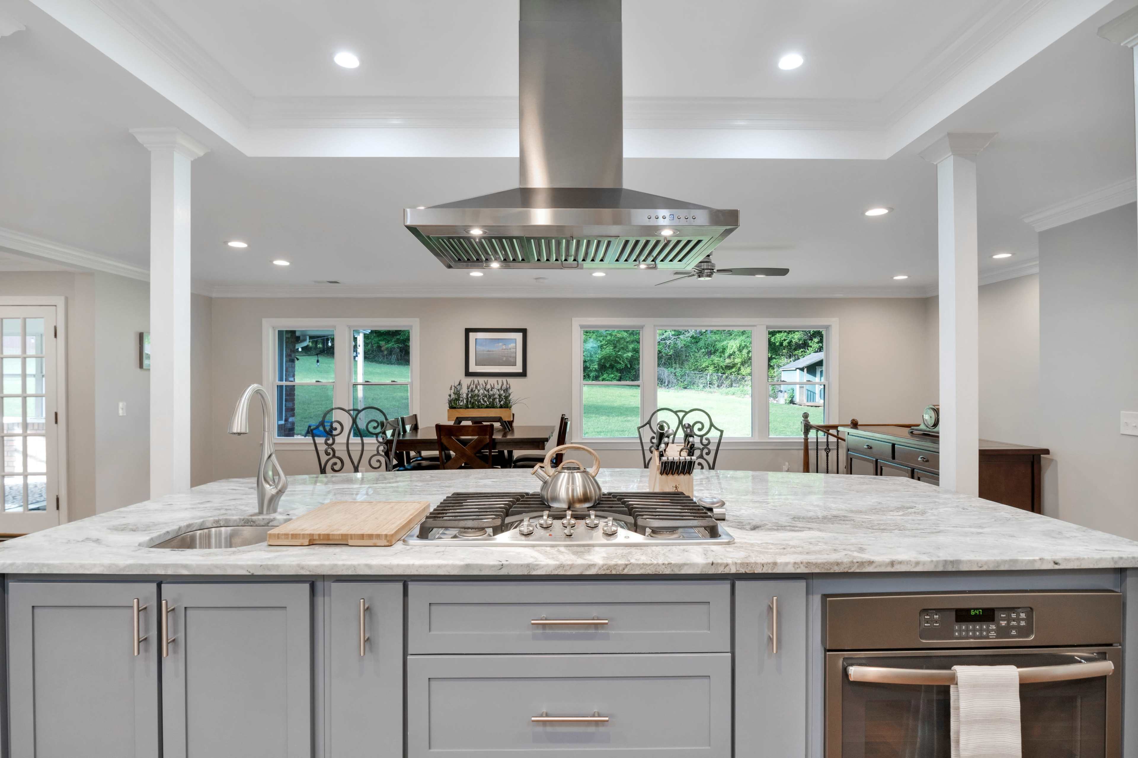 A modern kitchen features a central island with a stovetop and a hood, overlooking a dining area with large windows and a view of the outdoors.