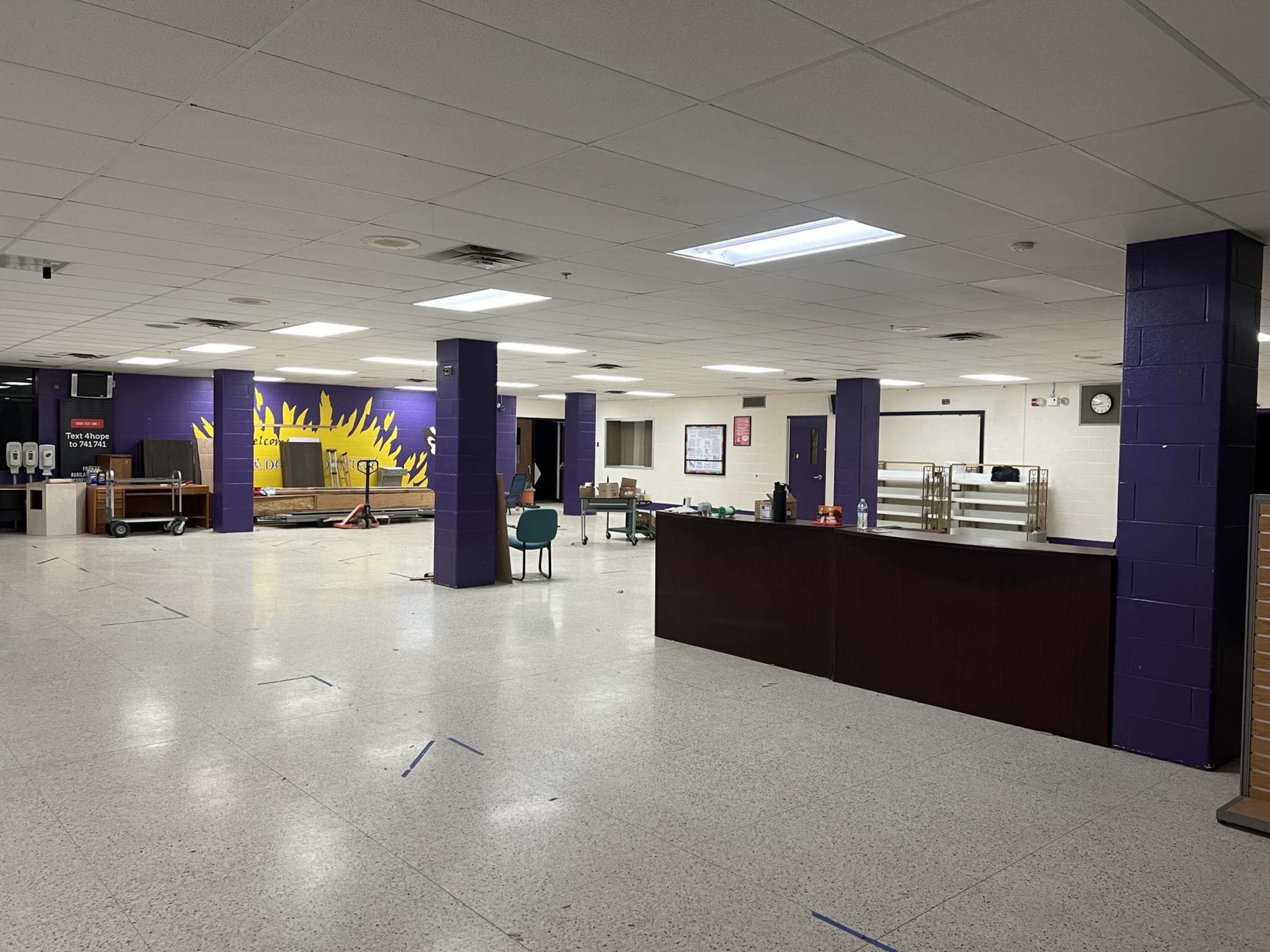 The image shows an empty cafeteria with a painted sun mural on one wall and tables stacked against the side.