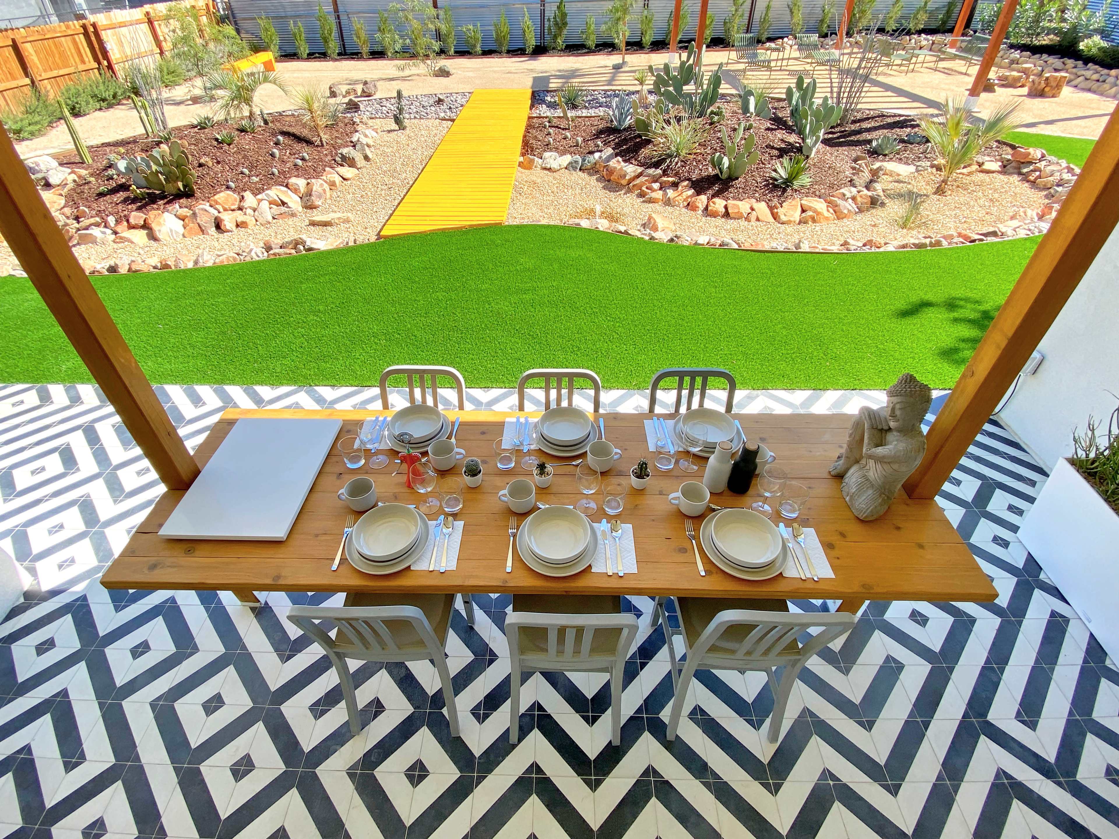 A long wooden dining table is set for a meal under a covered patio, with an outdoor garden featuring artificial turf and landscaped beds in the background.