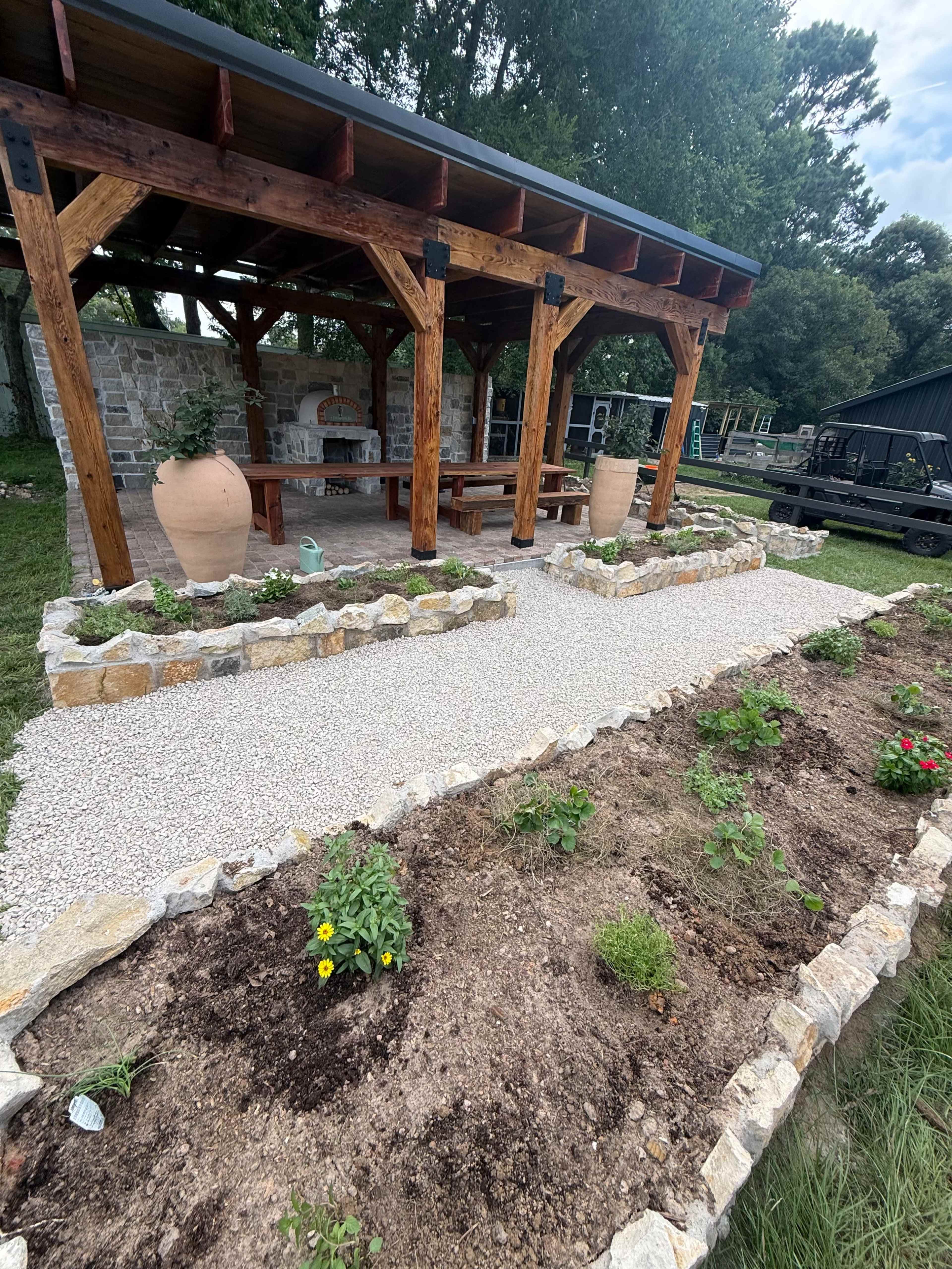 The image shows a spacious outdoor pavilion made of wood, featuring stone planters and a gravel pathway lined with freshly planted flowers and plants.