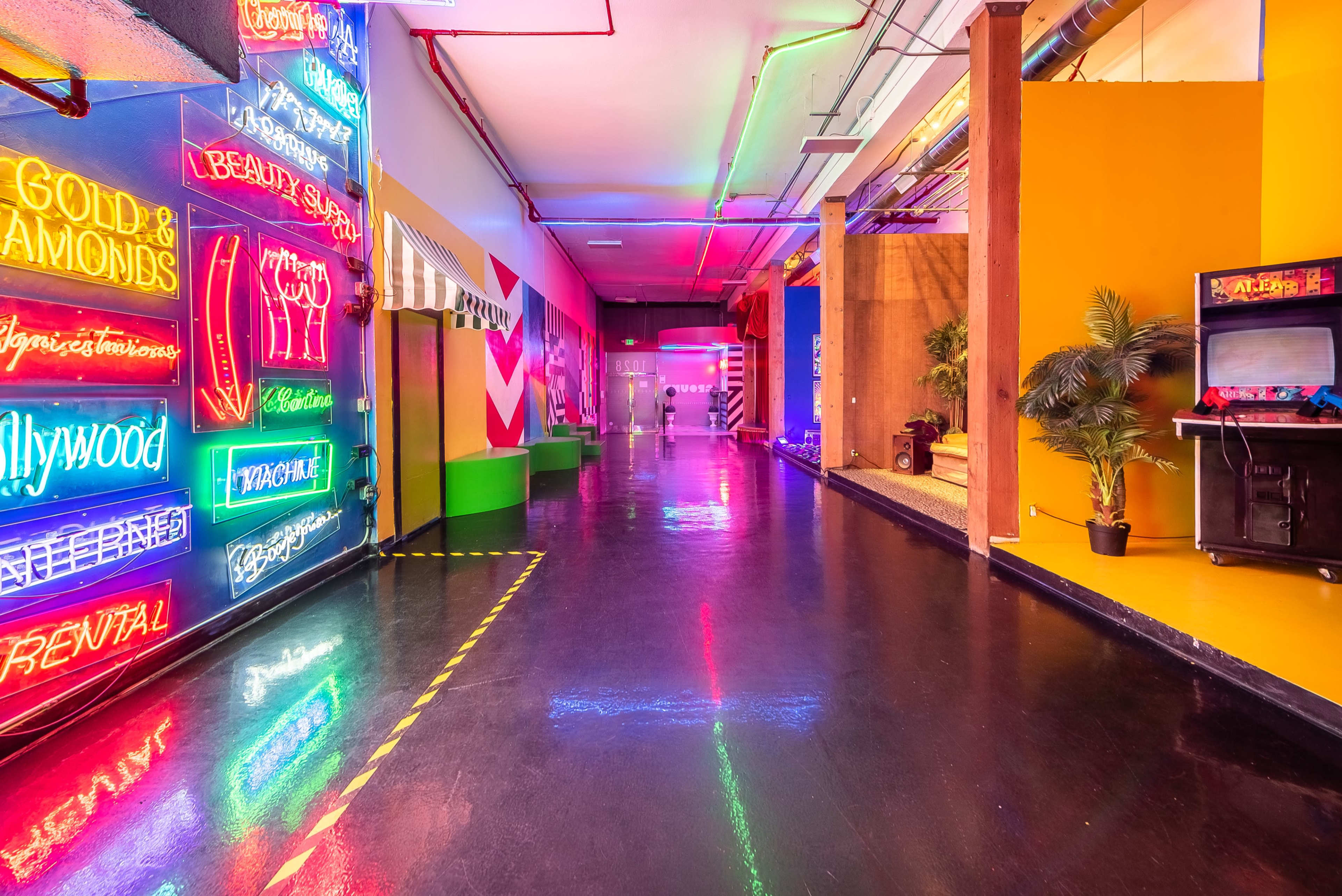 A brightly lit hallway with colorful neon signs and vivid wall colors, lined with reflective flooring and decorations, including a small plant and an arcade machine.