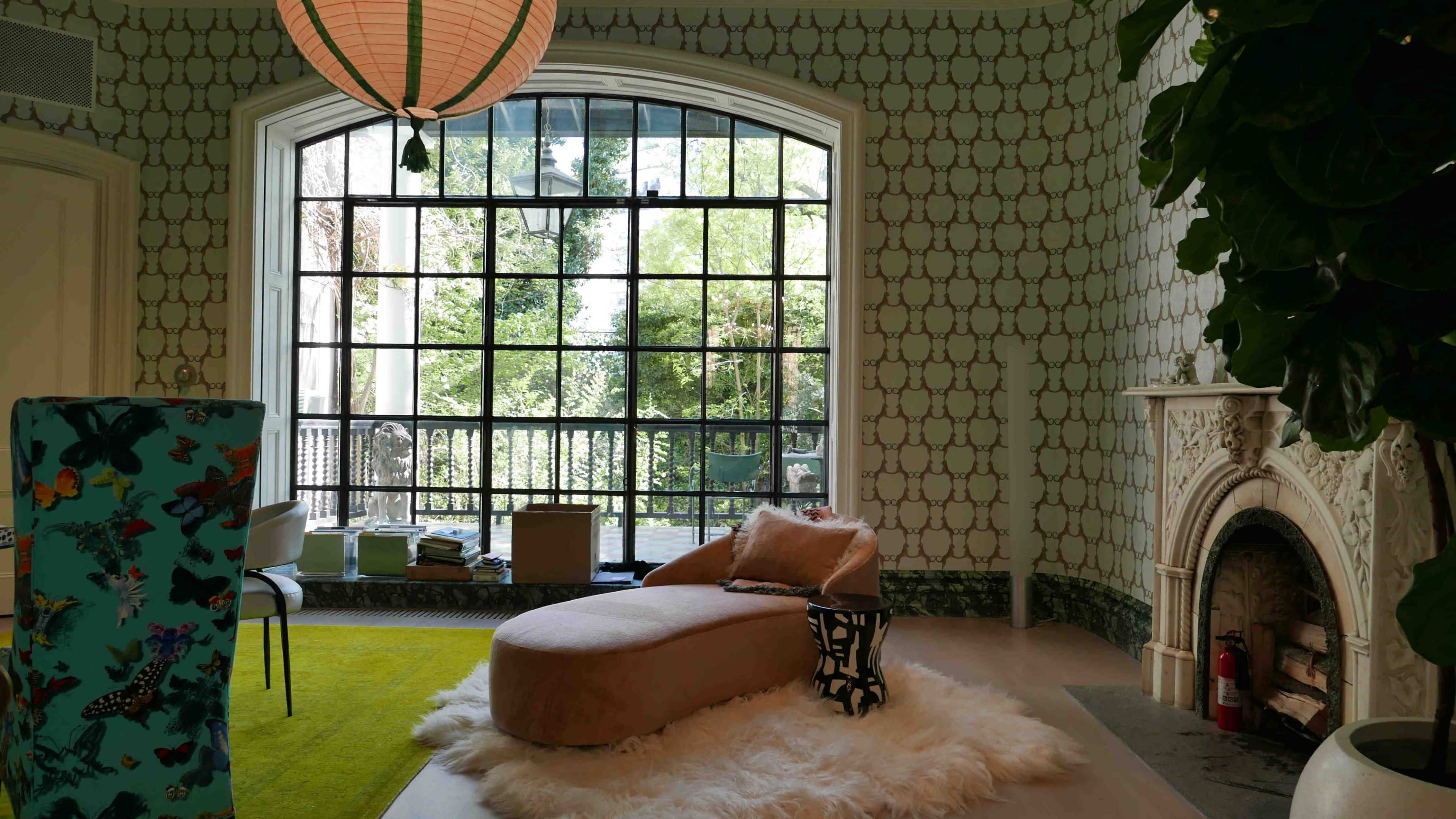 A stylish room features a large arched window overlooking greenery, with a pink chaise lounge on a white rug, and patterned wallpaper adorning the walls.