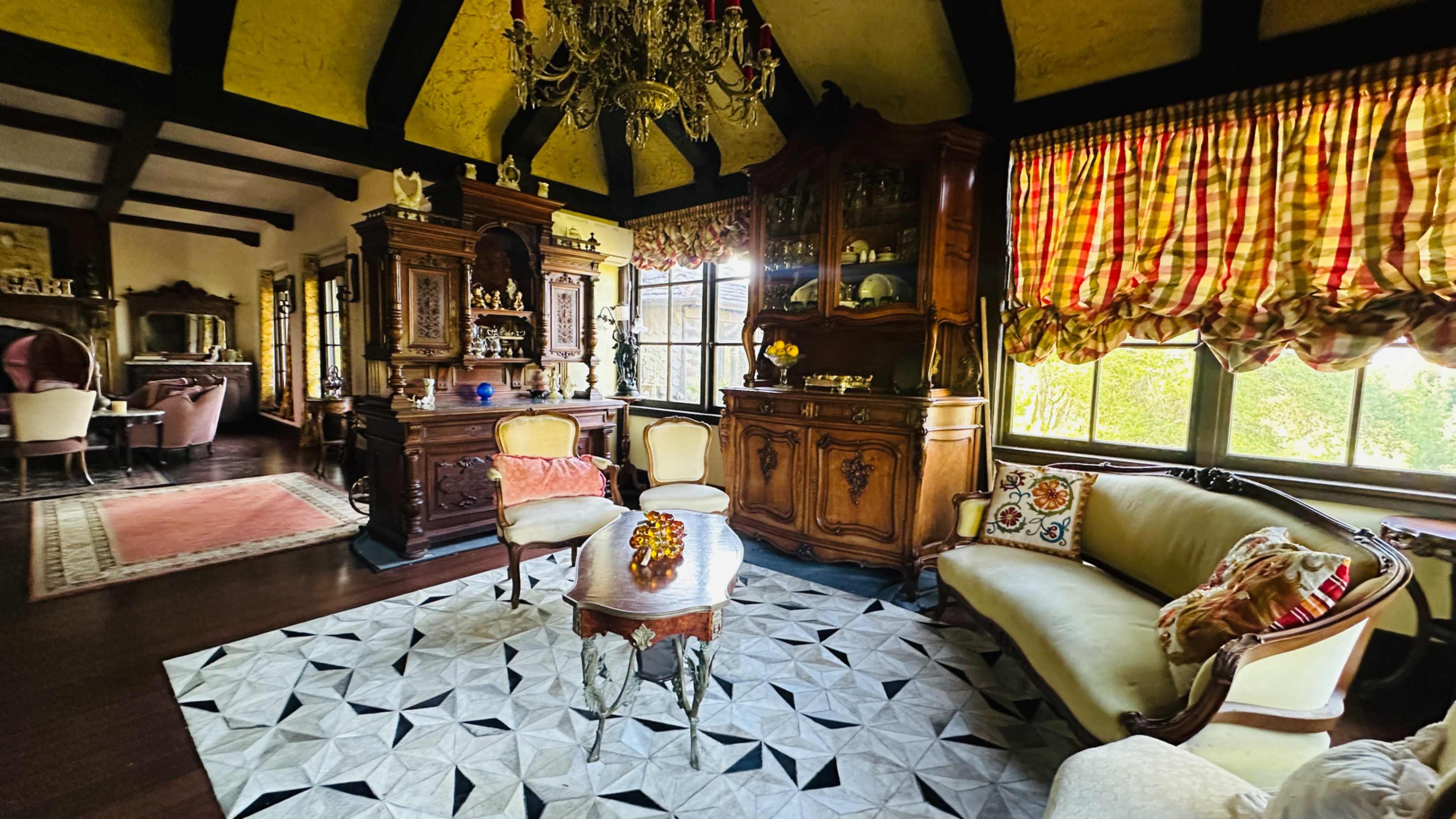 The image shows a vintage living room with a chandelier, ornate wooden furniture, and a patterned rug.