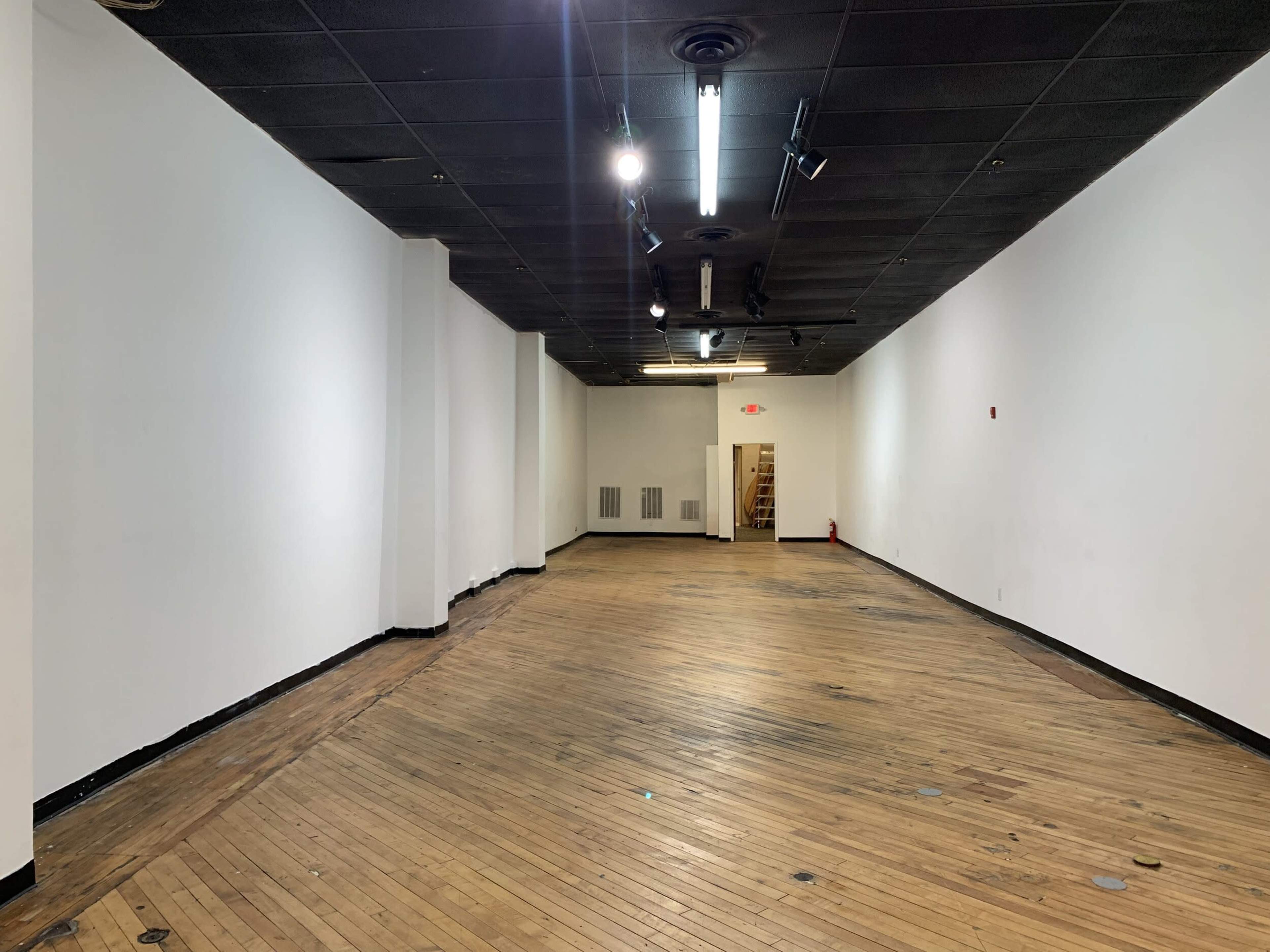 A large, empty interior space with wooden floors and plain white walls, featuring track lighting on the ceiling.